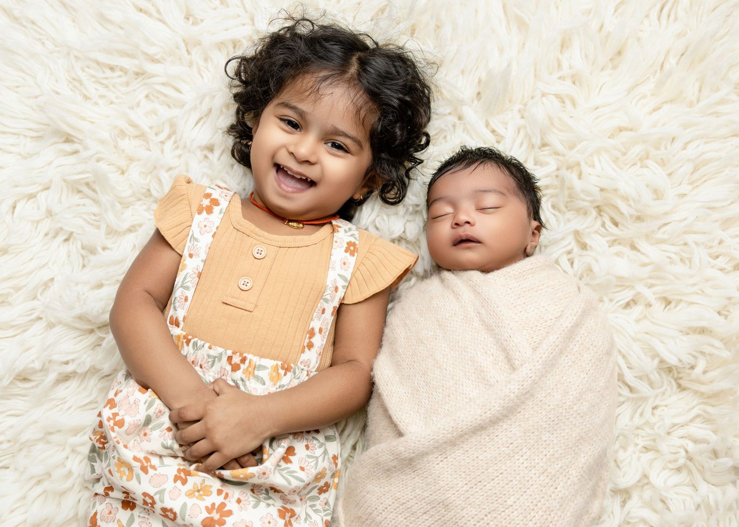 A smiling toddler girl poses beside a sleeping swaddled newborn baby on a fluffy white rug.