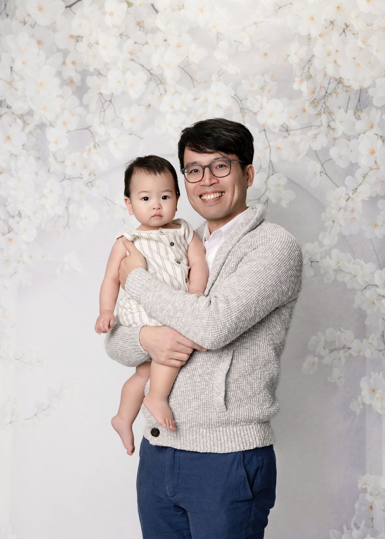 Smiling Asian father holding baby in striped outfit against white floral studio backdrop.