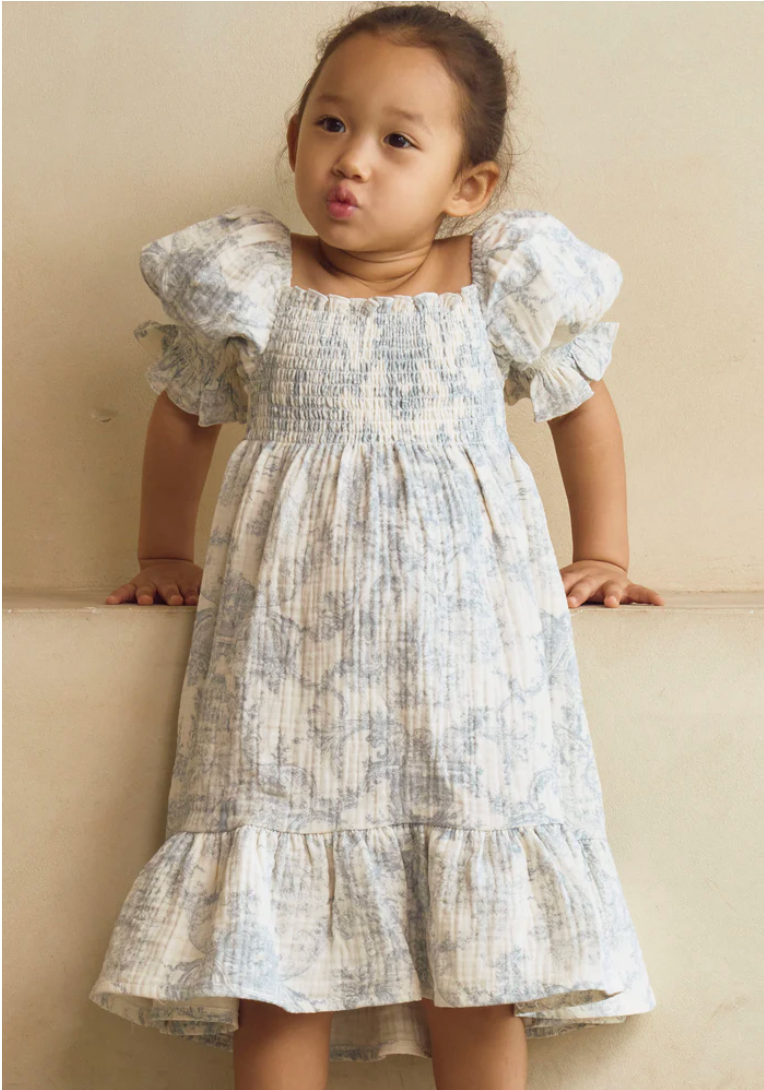 Toddler girl wearing a white floral smocked dress with ruffle sleeves and tiered skirt.