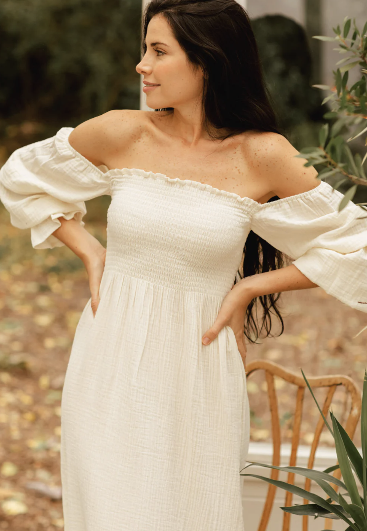 Woman wearing an off-shoulder white smocked midi dress with puff sleeves, posed outdoors in a garden setting.
