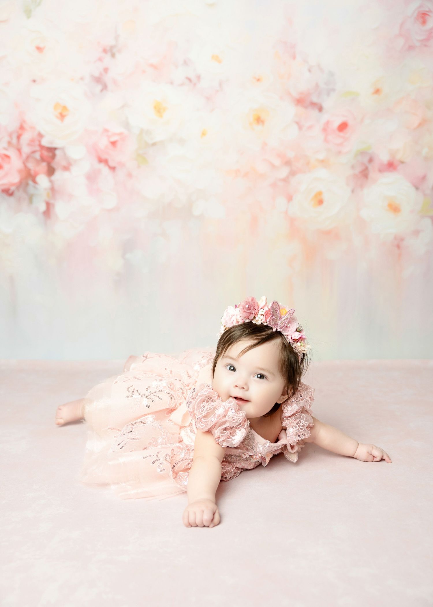 Cute baby in pink lace dress and floral headband crawls on pink floor with floral backdrop.