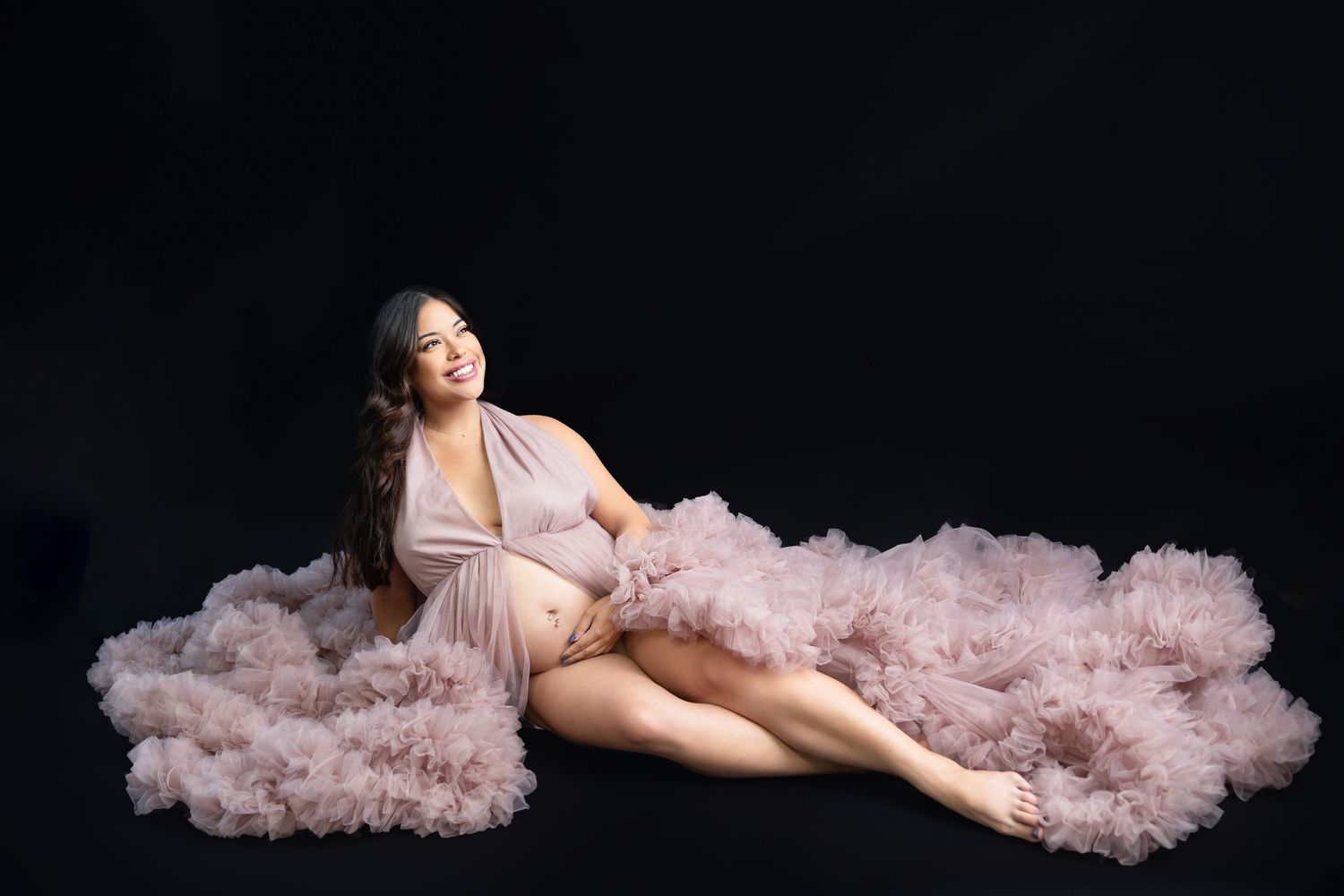 Smiling pregnant woman reclined in elegant pink ruffled tulle maternity dress on dark background.