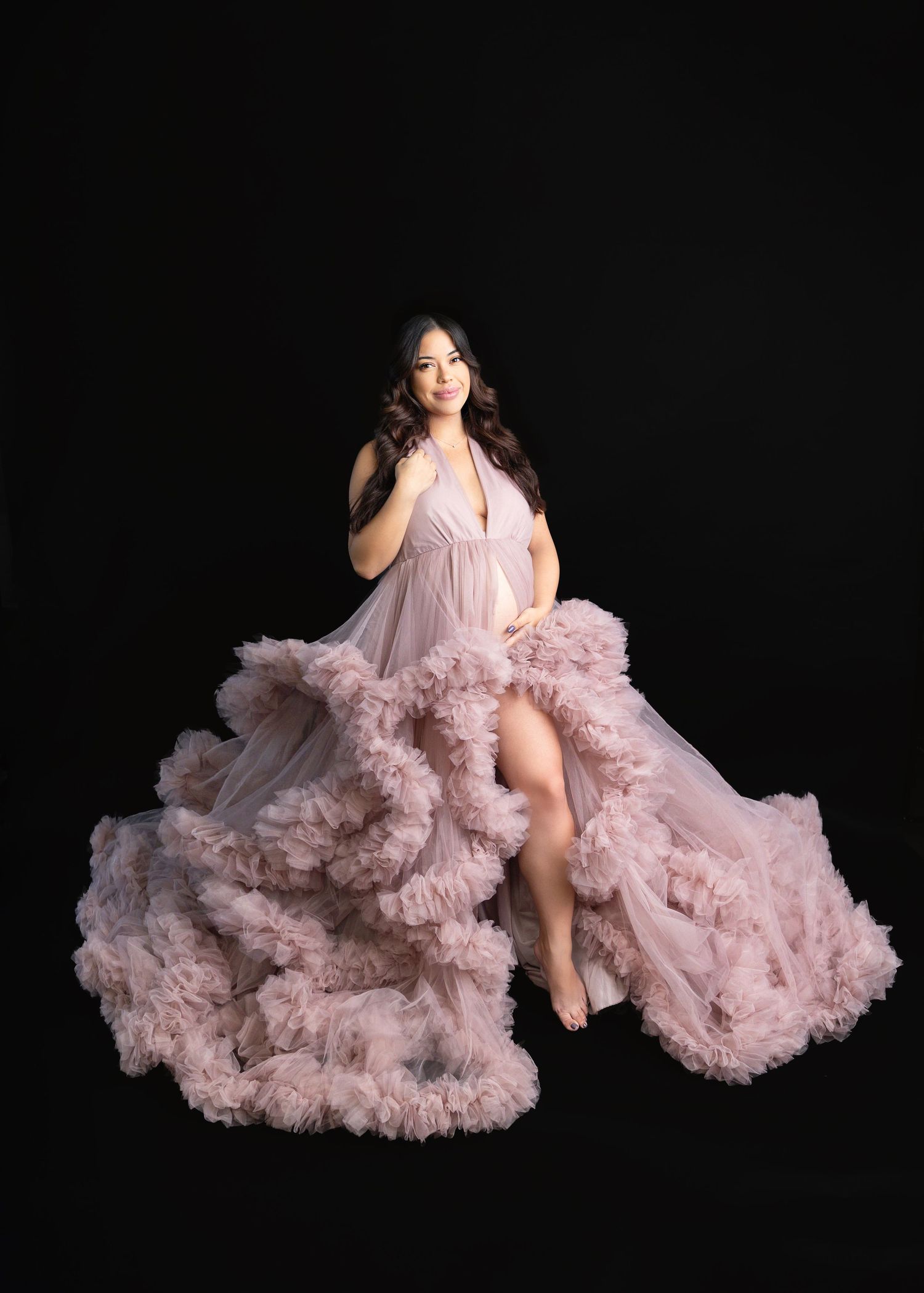 Pregnant woman in flowing pink ruffle tulle maternity gown posing elegantly against black background.