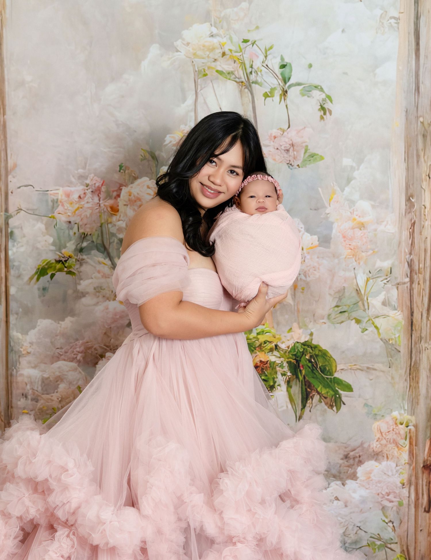 Woman in pink tulle gown holding newborn baby, posed before a soft floral backdrop in a dreamy portrait.