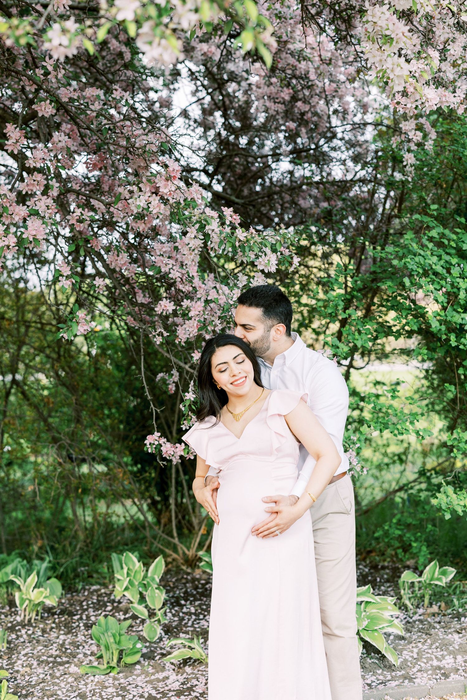Couple embracing under blooming pink spring tree, pregnant woman in blush dress, maternity photo session outdoors.