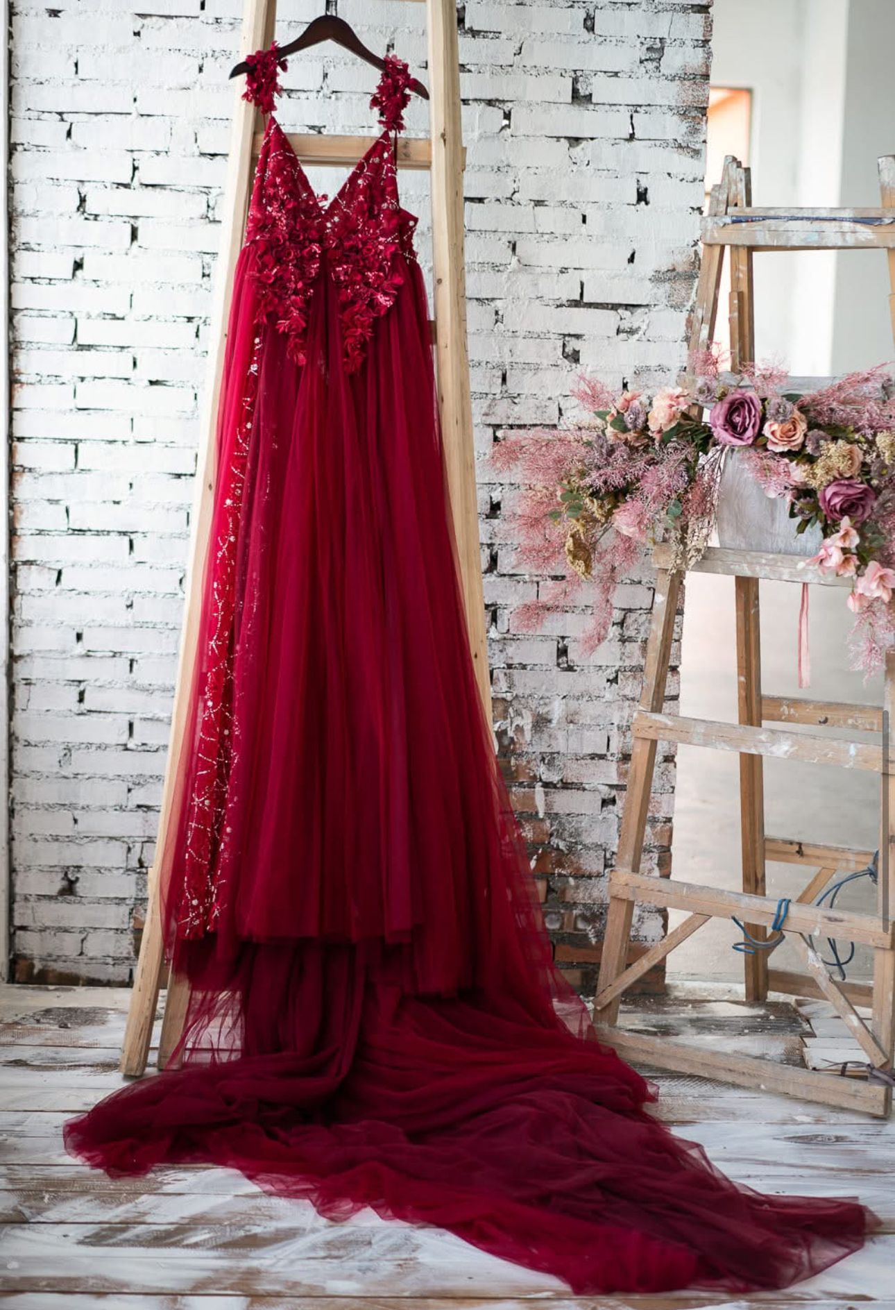 Elegant burgundy tulle ball gown with floral lace bodice hanging on a wooden ladder against a white brick wall.
