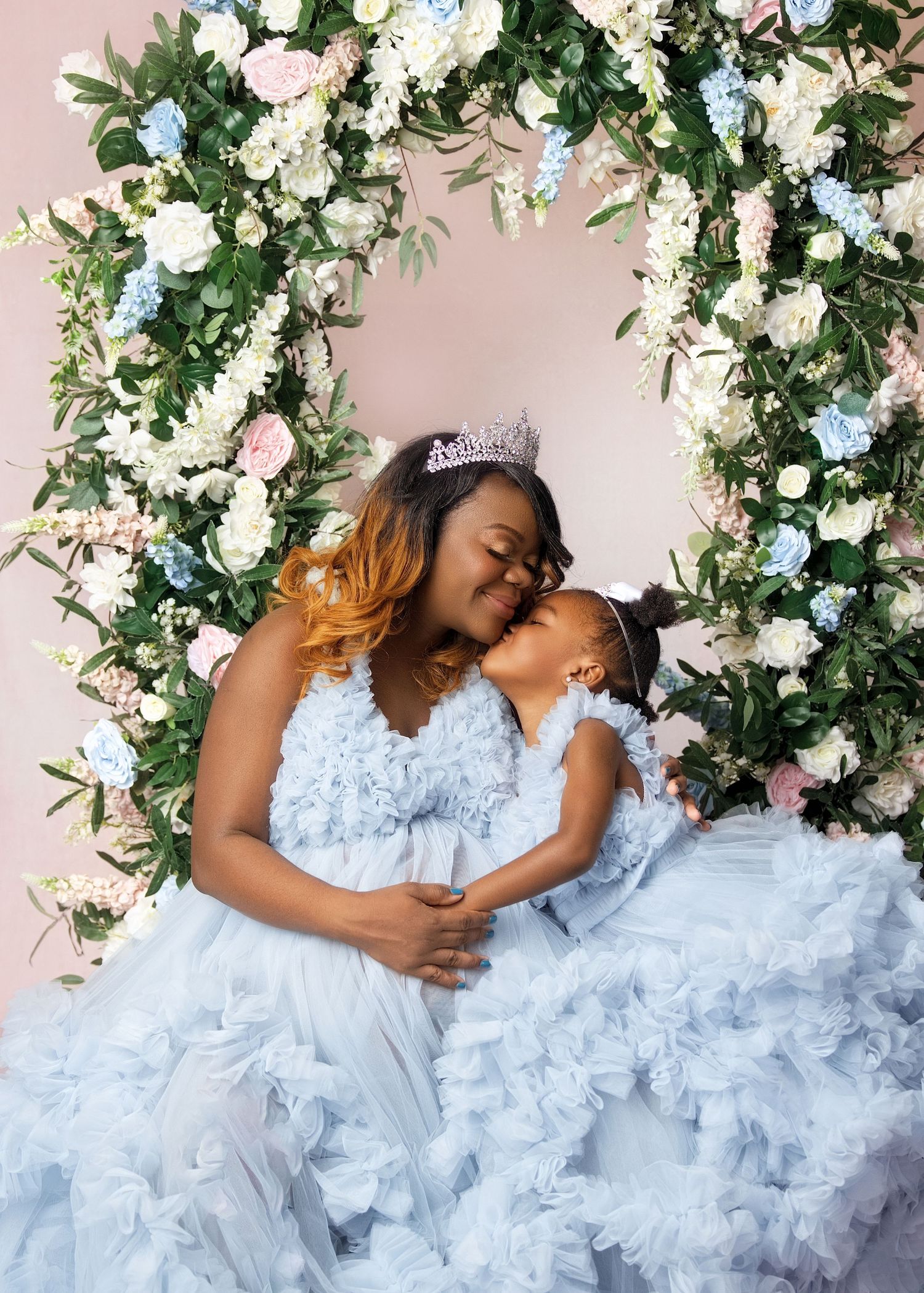 A mother and daughter in matching light blue ruffle gowns share a tender kiss surrounded by a floral arch.