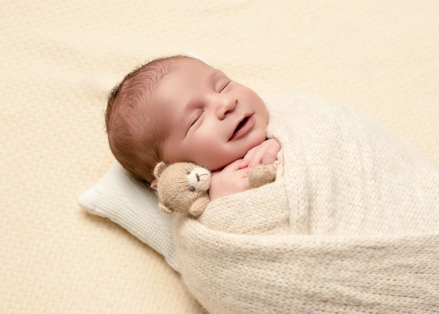 Sleeping newborn baby swaddled in cream wrap holding a tiny teddy bear with a gentle smile.