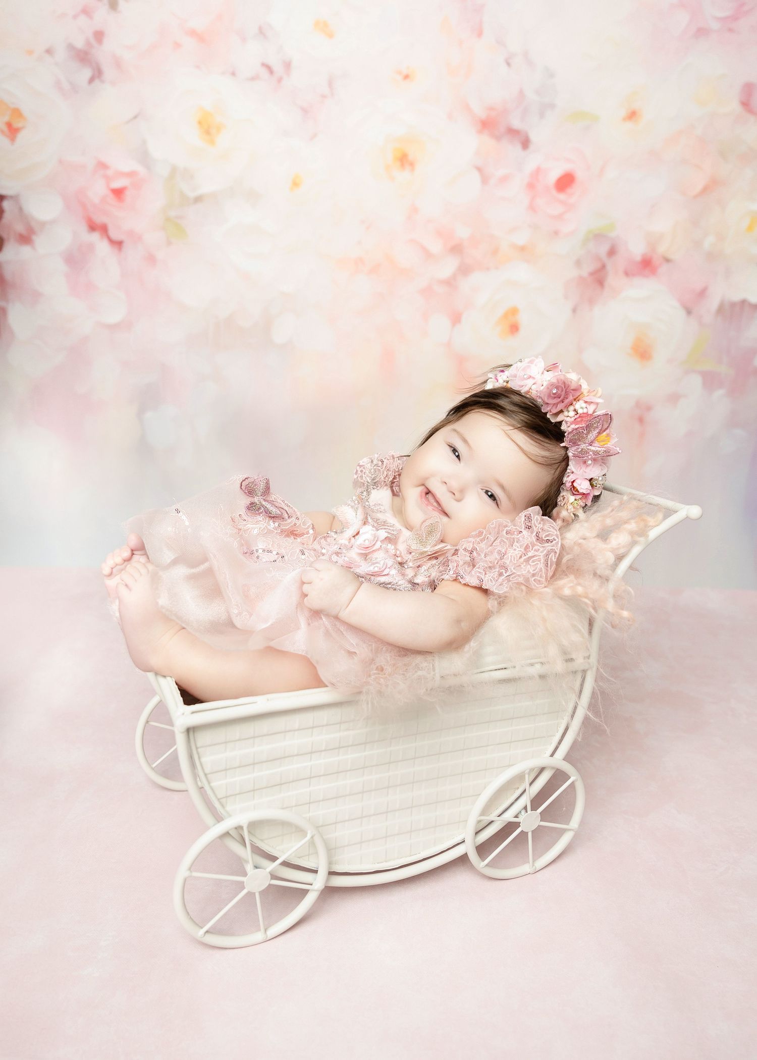 Joyful baby in pink lace romper smiles inside a vintage white miniature baby carriage.