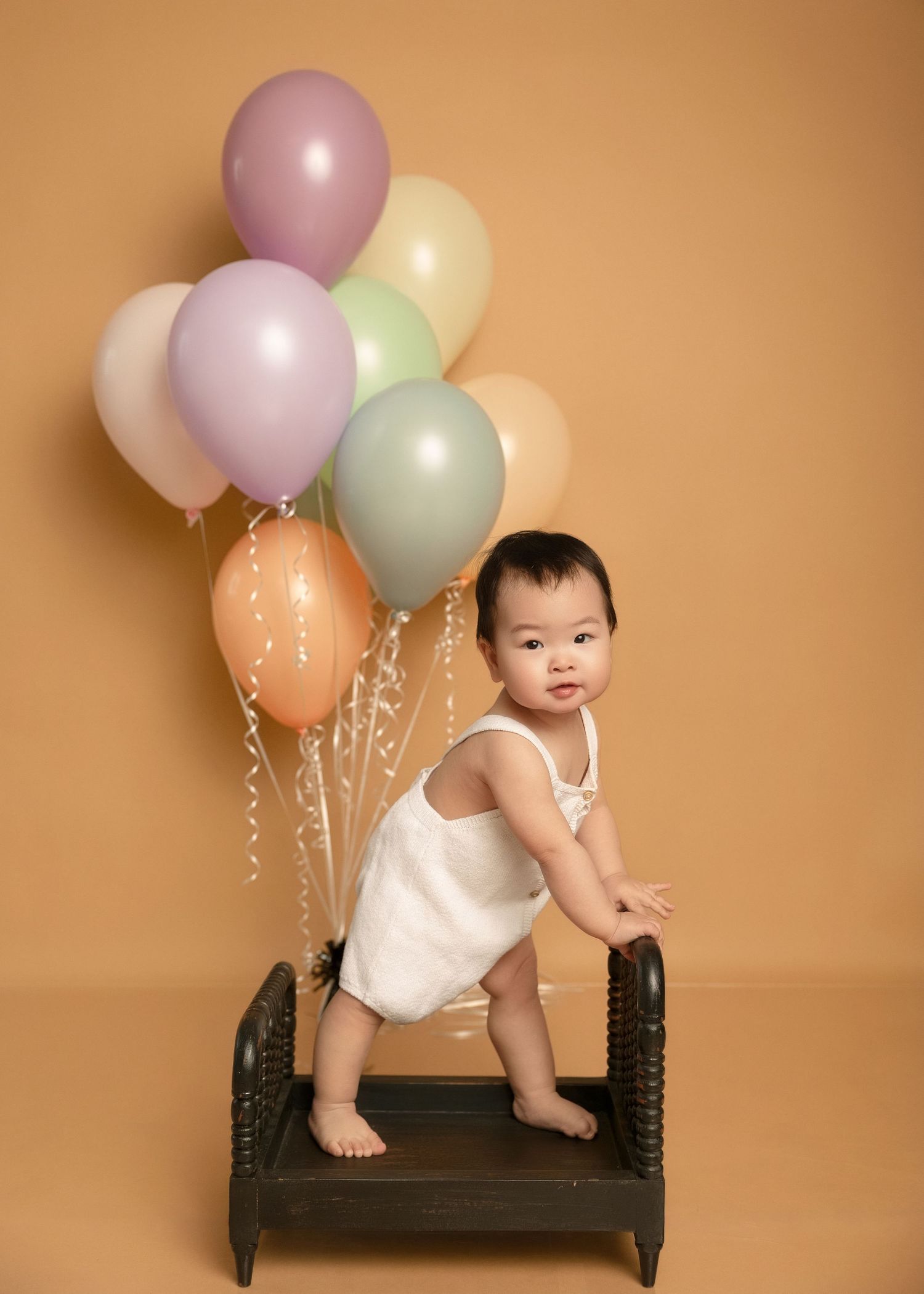 Baby in white romper standing on small bench with colorful pastel balloons on warm beige background.