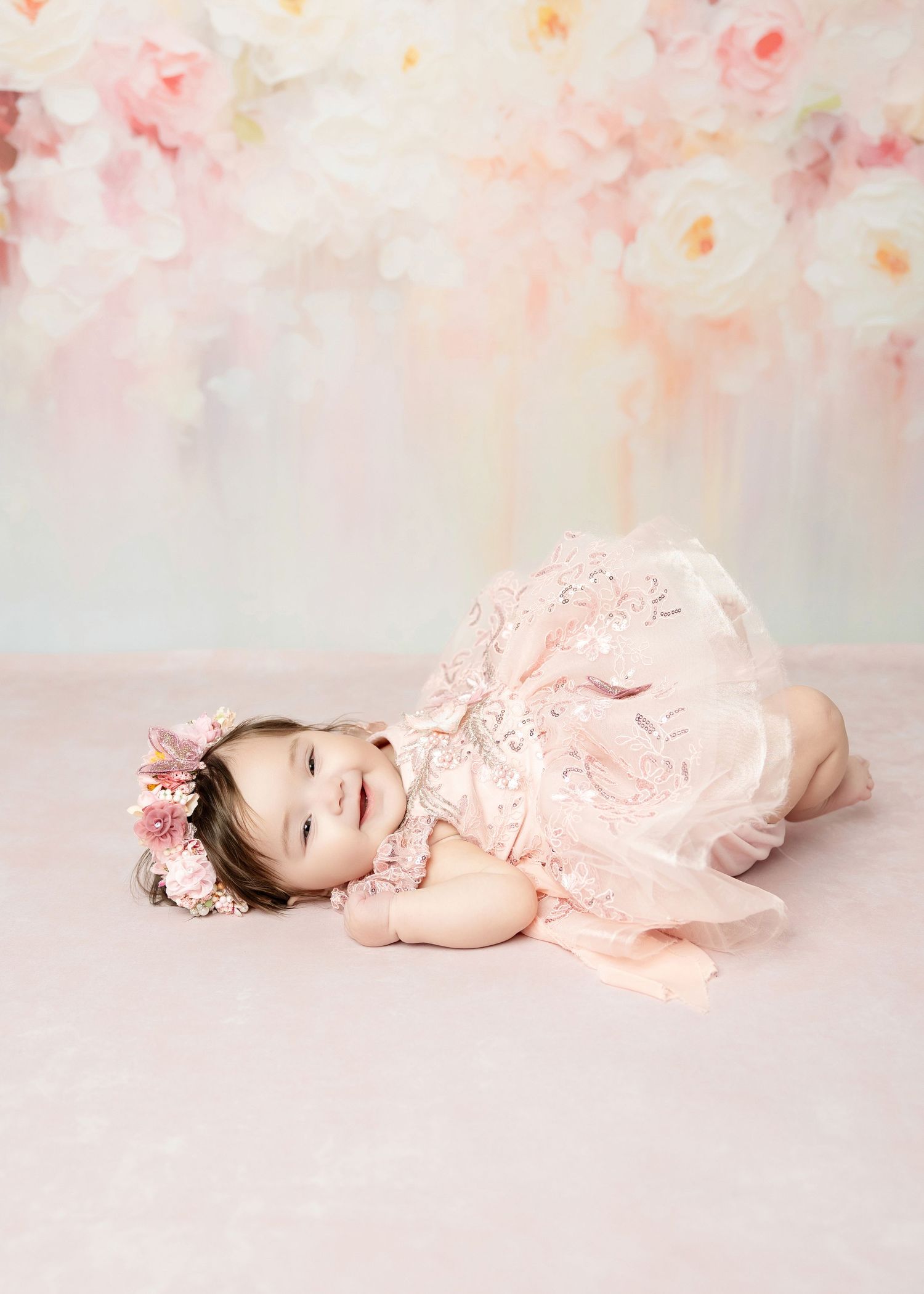 Smiling baby girl in pink lace romper lies on soft pink floor with floral backdrop.