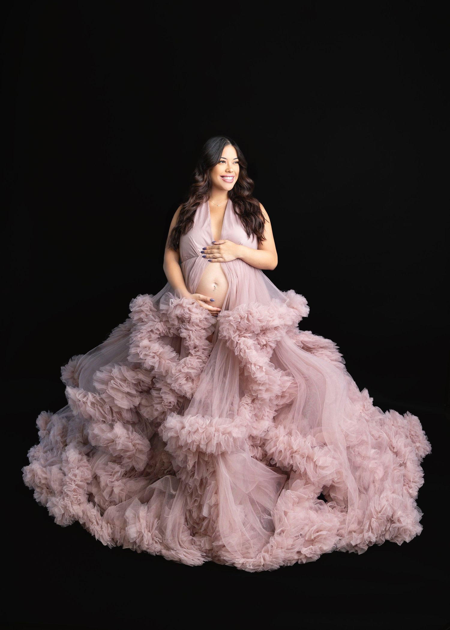 Smiling pregnant woman in dramatic pink ruffled tulle gown posing against solid black background.