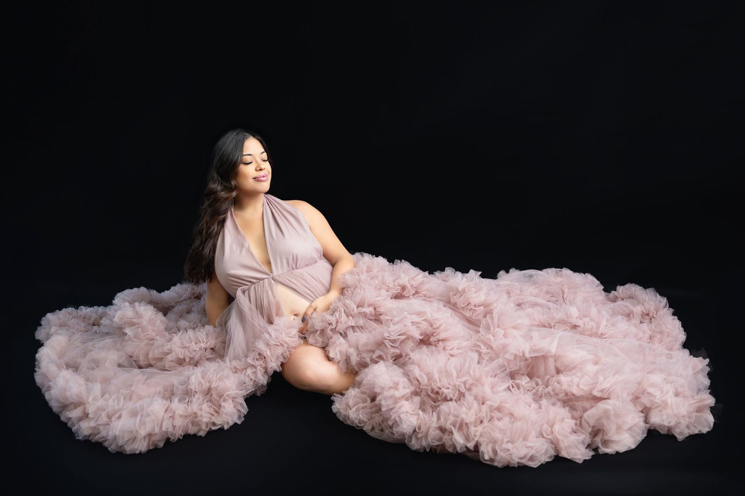 Pregnant woman sitting gracefully in voluminous blush pink tulle gown on black studio backdrop.