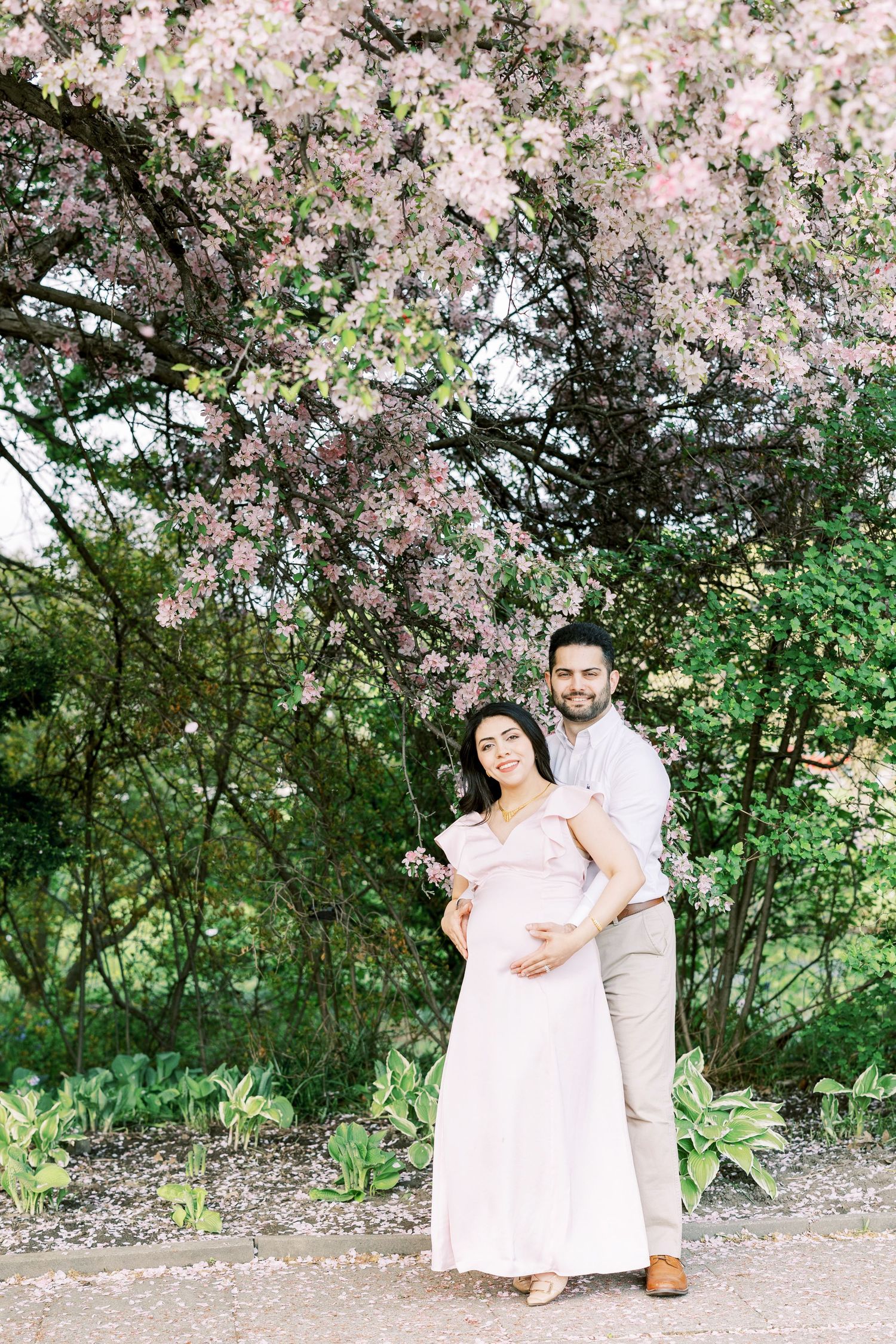 Expecting couple posing under flowering pink tree, man holding pregnant woman in blush maternity dress outdoors.