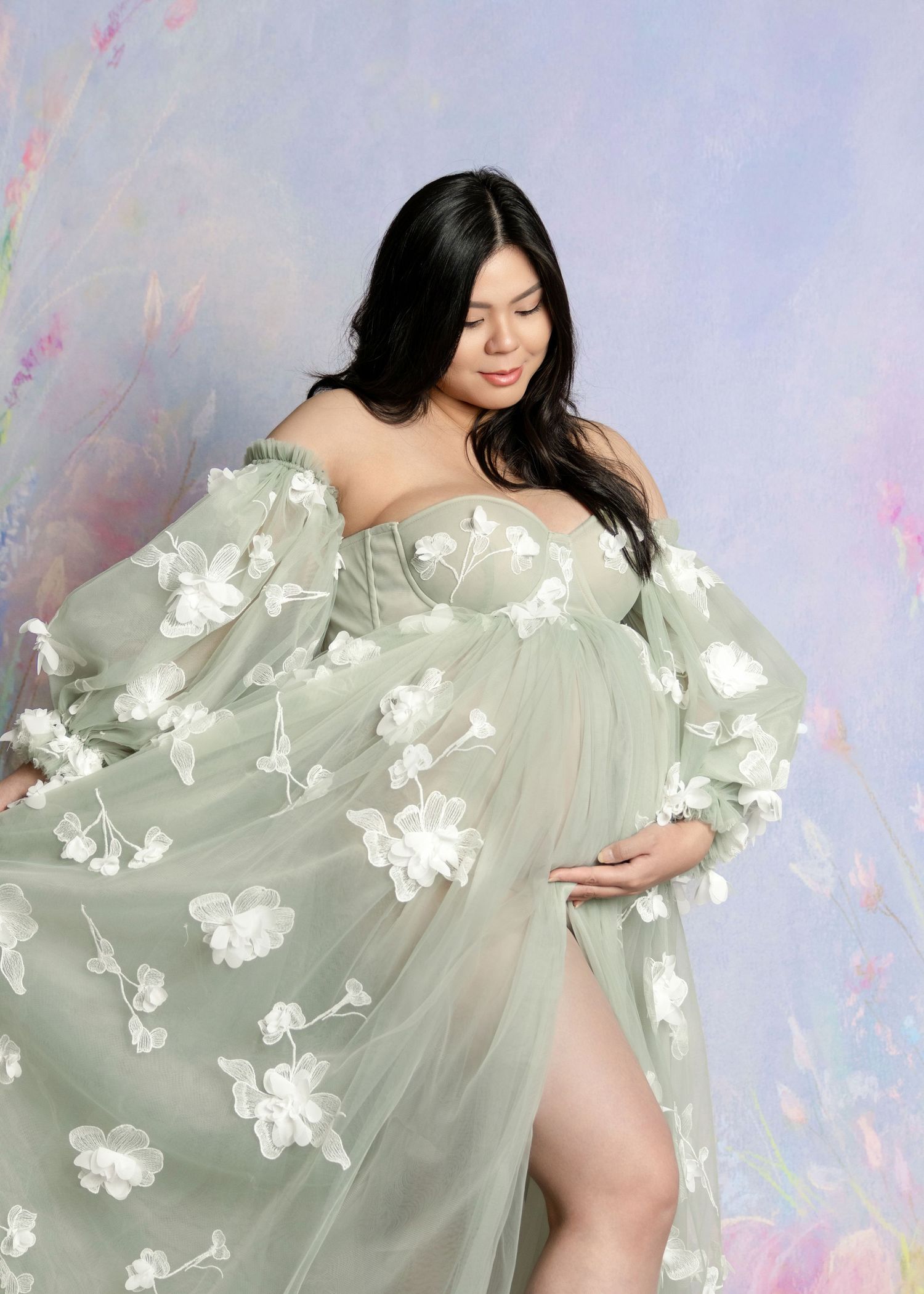 Pregnant woman in sage green floral tulle maternity gown posing gracefully against a soft pastel floral backdrop.