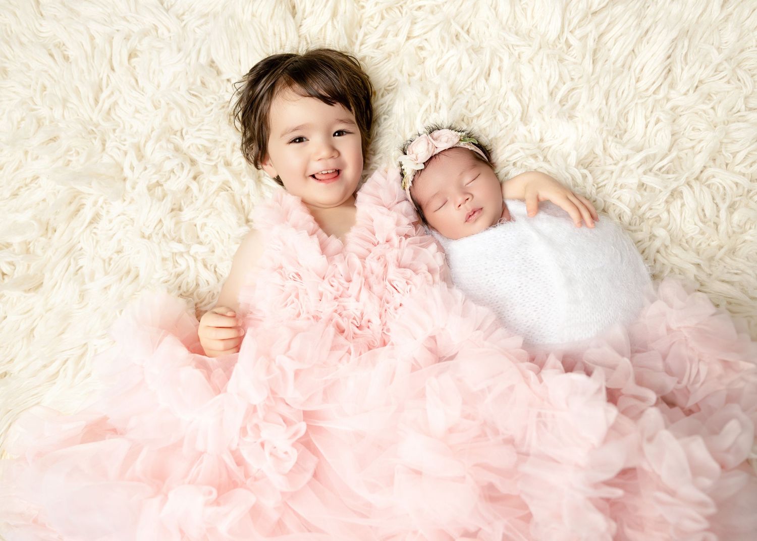 Smiling toddler girl in pink dress posing with her sleeping swaddled newborn sibling on white rug.