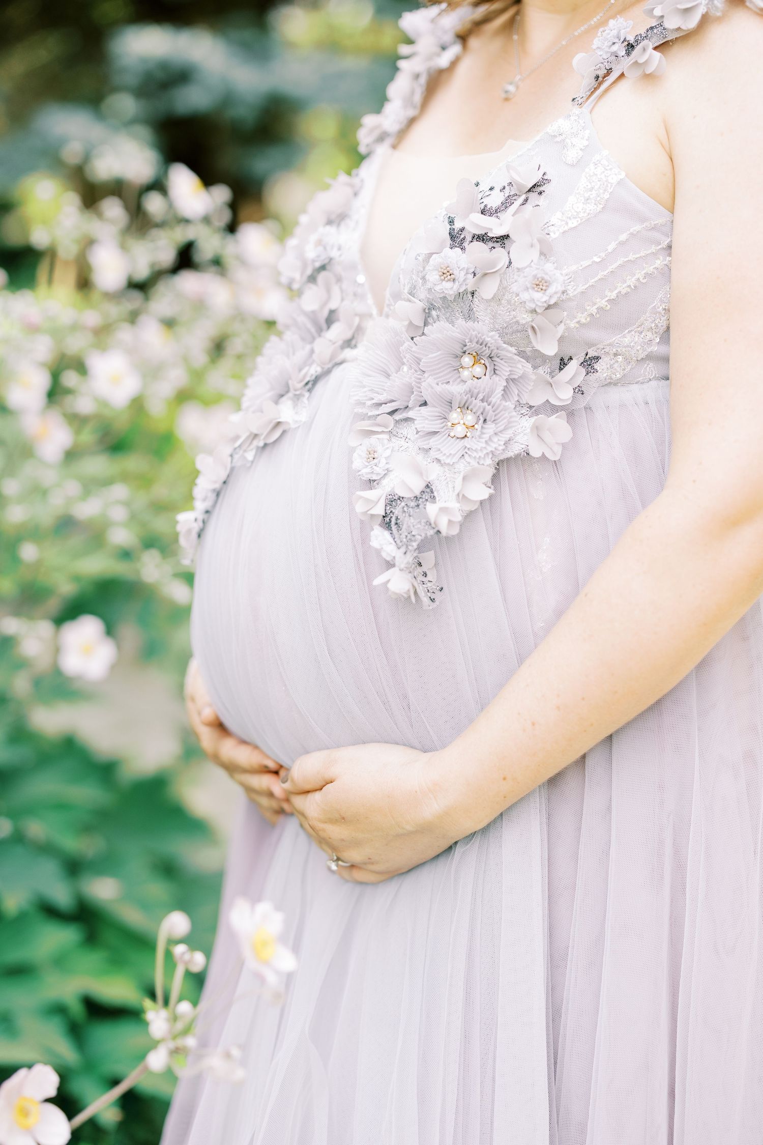 Close-up of pregnant belly in lavender floral applique maternity gown surrounded by white garden flowers.
