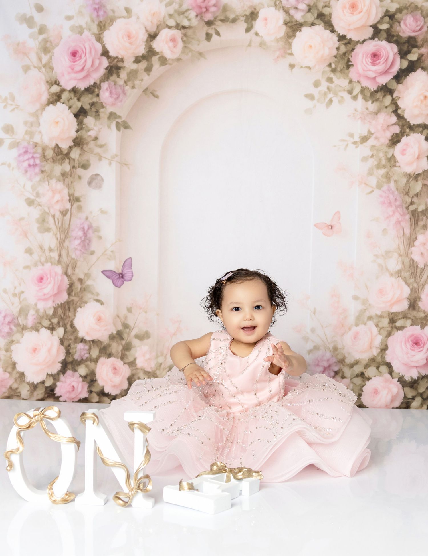 Baby girl in pink dress sits before floral arch backdrop with ONE letters for first birthday photo.