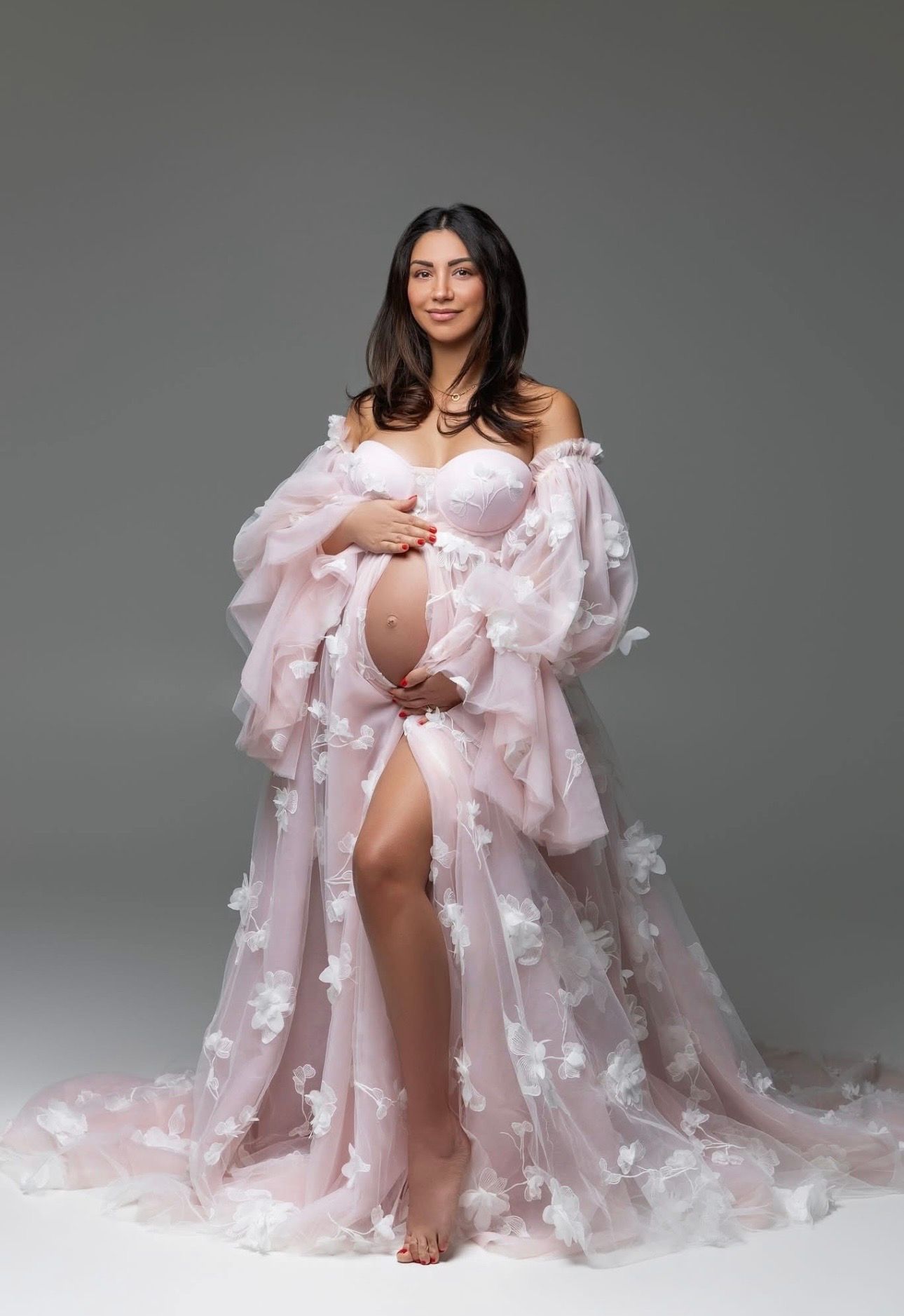 Pregnant woman in a pink floral tulle maternity gown posing gracefully in a studio setting.