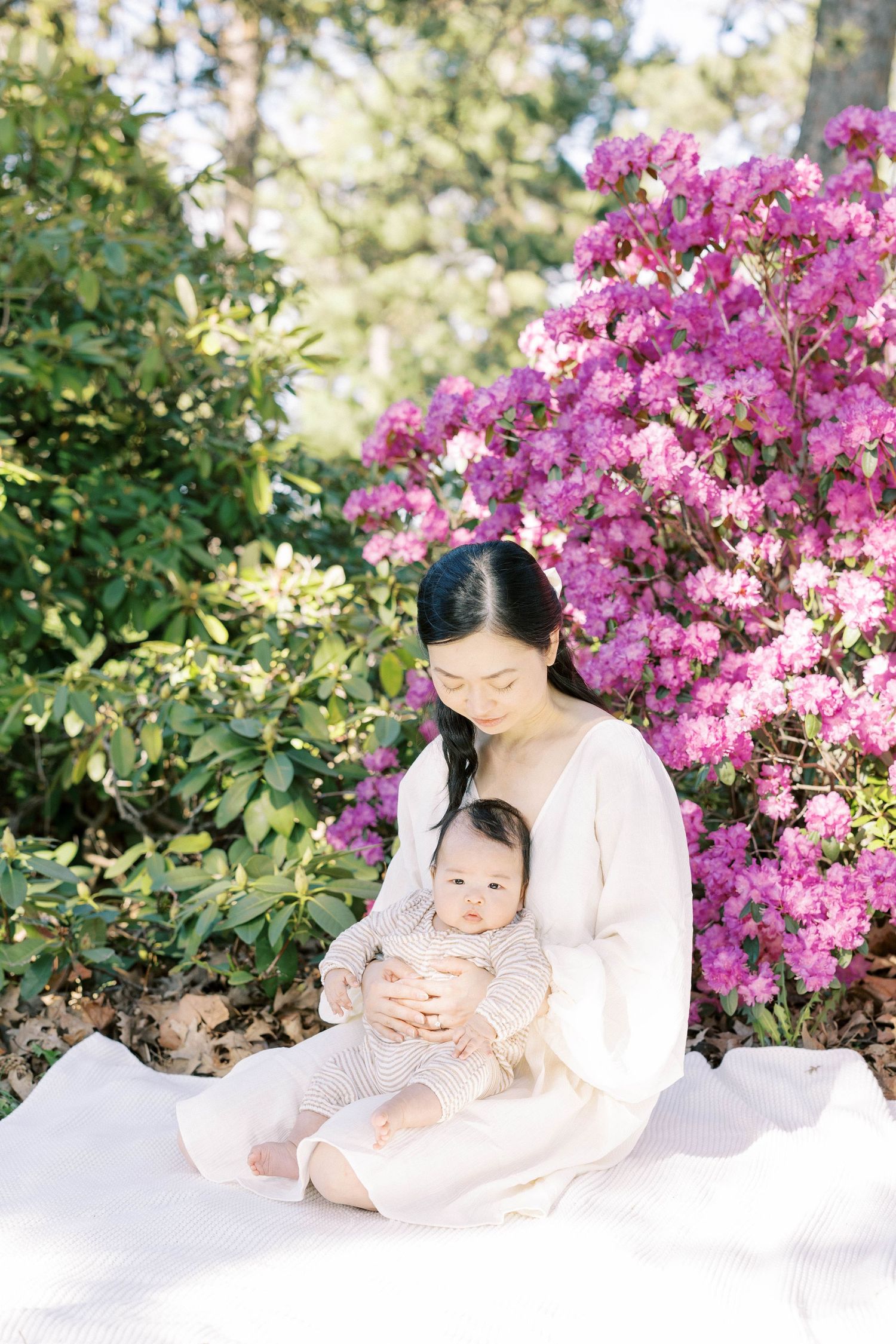 Serene Asian mother in white dress sits on blanket cradling infant before lush pink flowering rhododendrons.