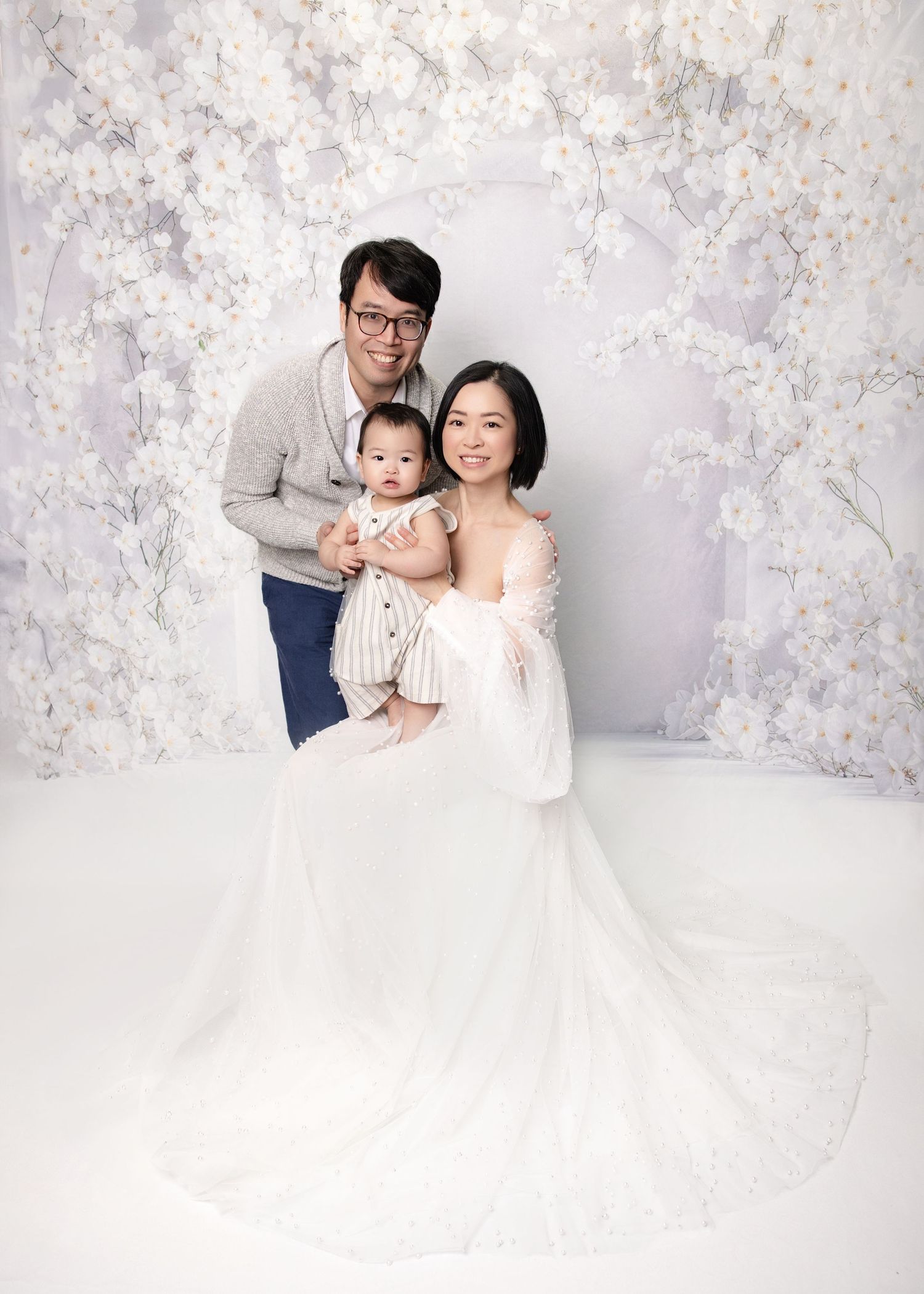 Asian family portrait with mother in white gown holding baby, father posing on floral backdrop.