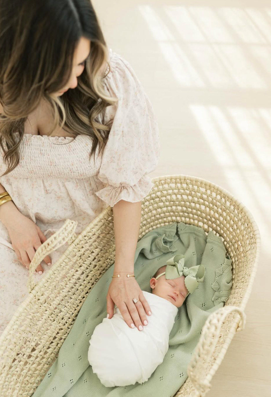 Mother gazing lovingly at swaddled newborn baby in woven Moses basket with sage green blanket and bow headband.