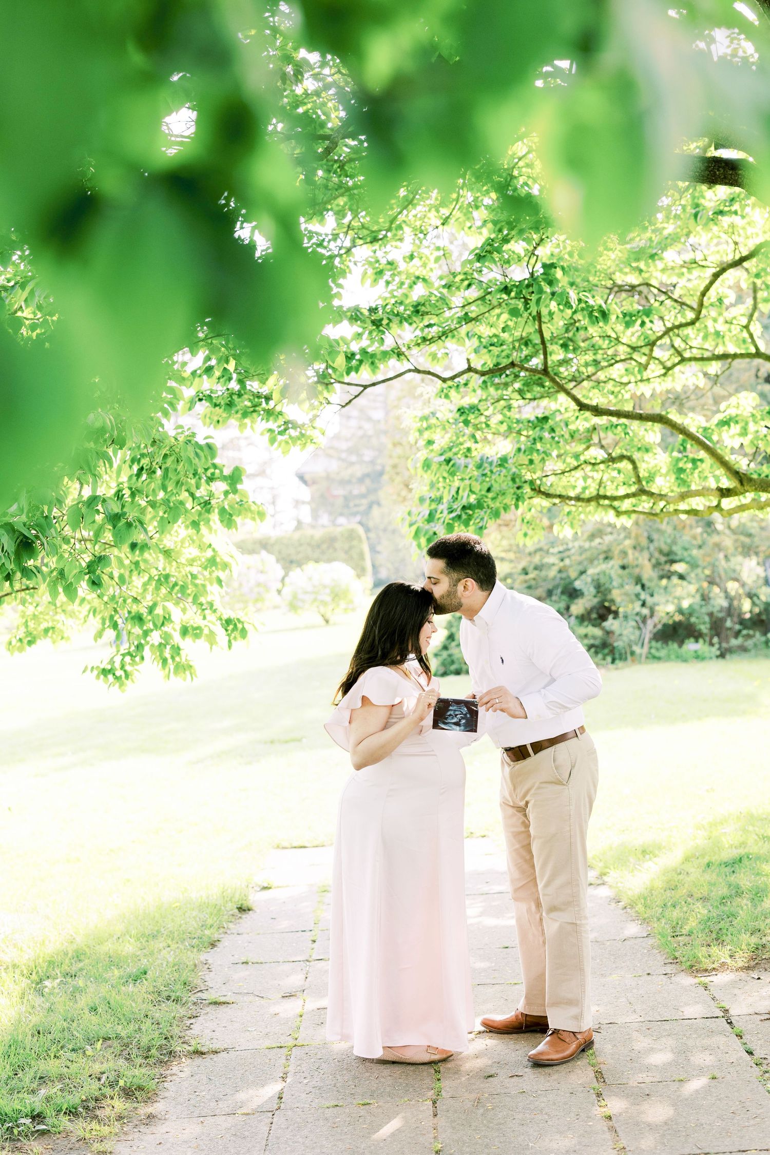 Expecting couple sharing tender moment holding ultrasound photo on garden path under lush green trees.