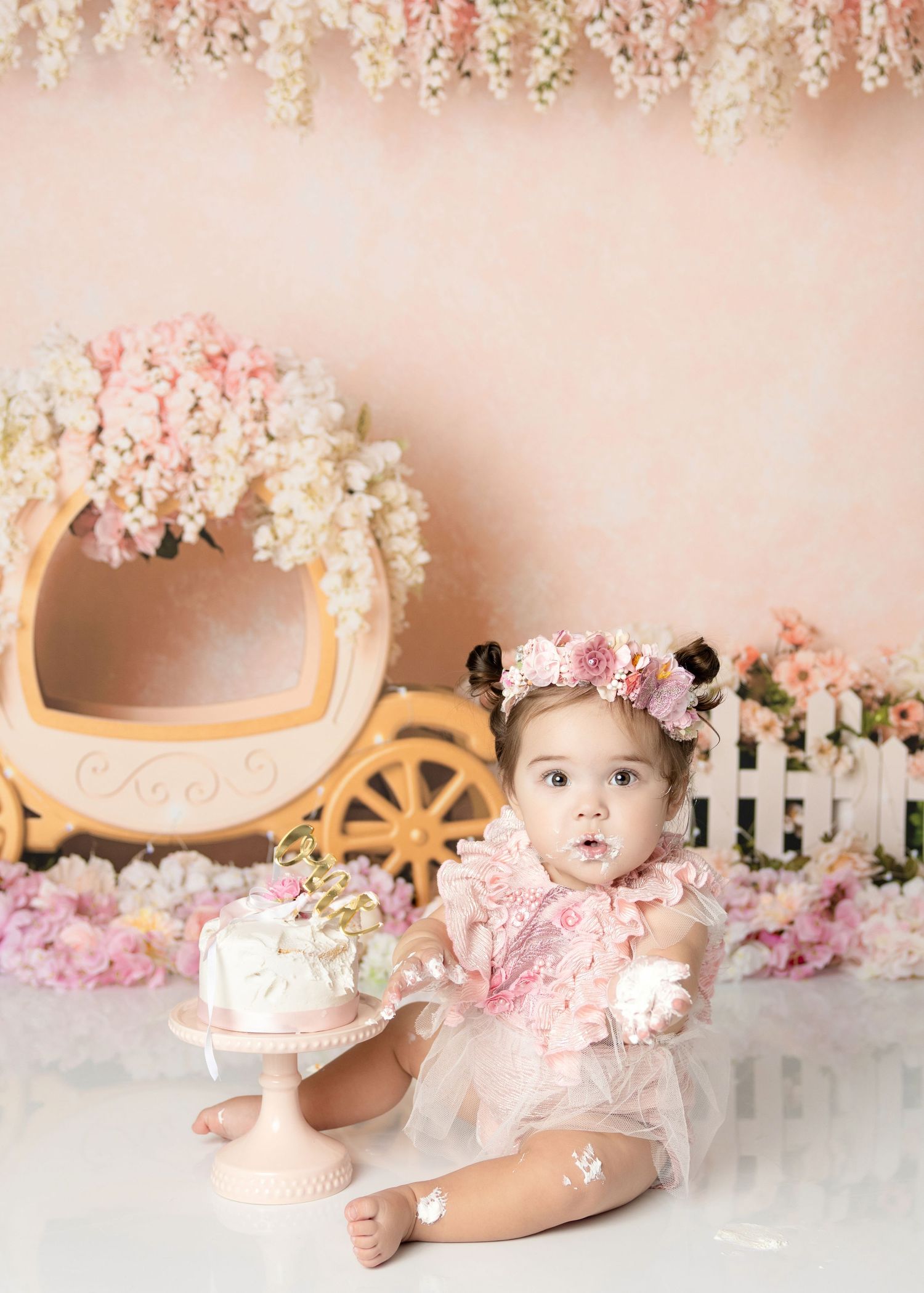 Surprised baby girl in pink floral romper sits beside first birthday cake with princess carriage backdrop.
