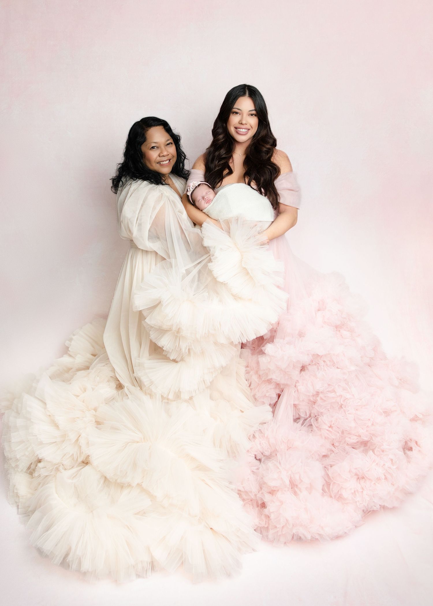 Two women in white and pink ruffled tulle gowns posing together with a newborn baby.