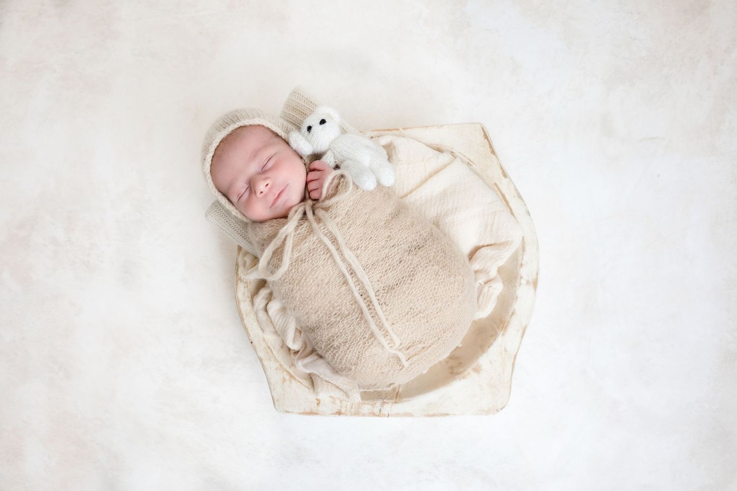 Sleeping newborn baby swaddled in cream wrap with teddy bear in wooden bowl on white fur.