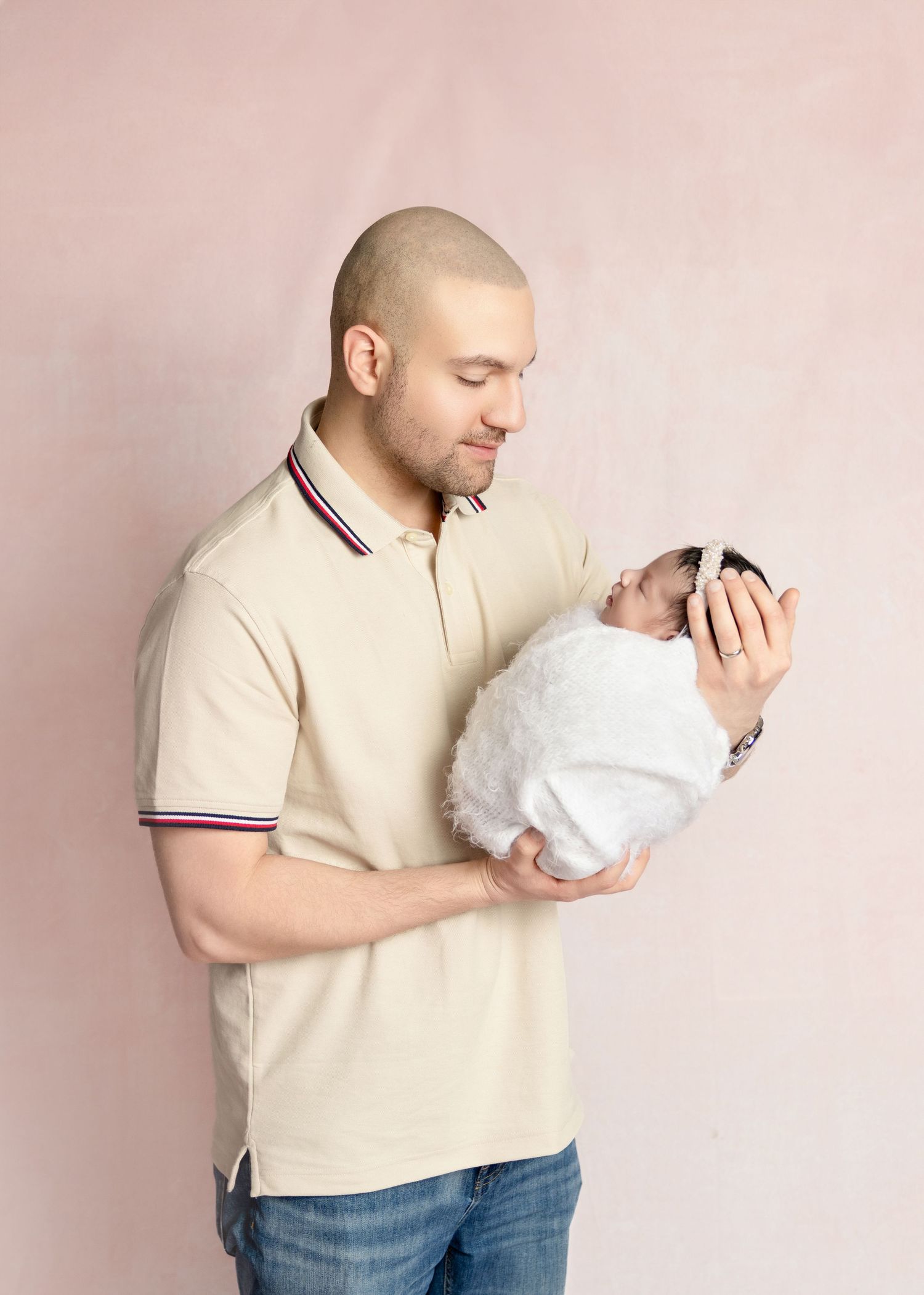 Father tenderly holds swaddled newborn baby during professional newborn photography session on pink backdrop.
