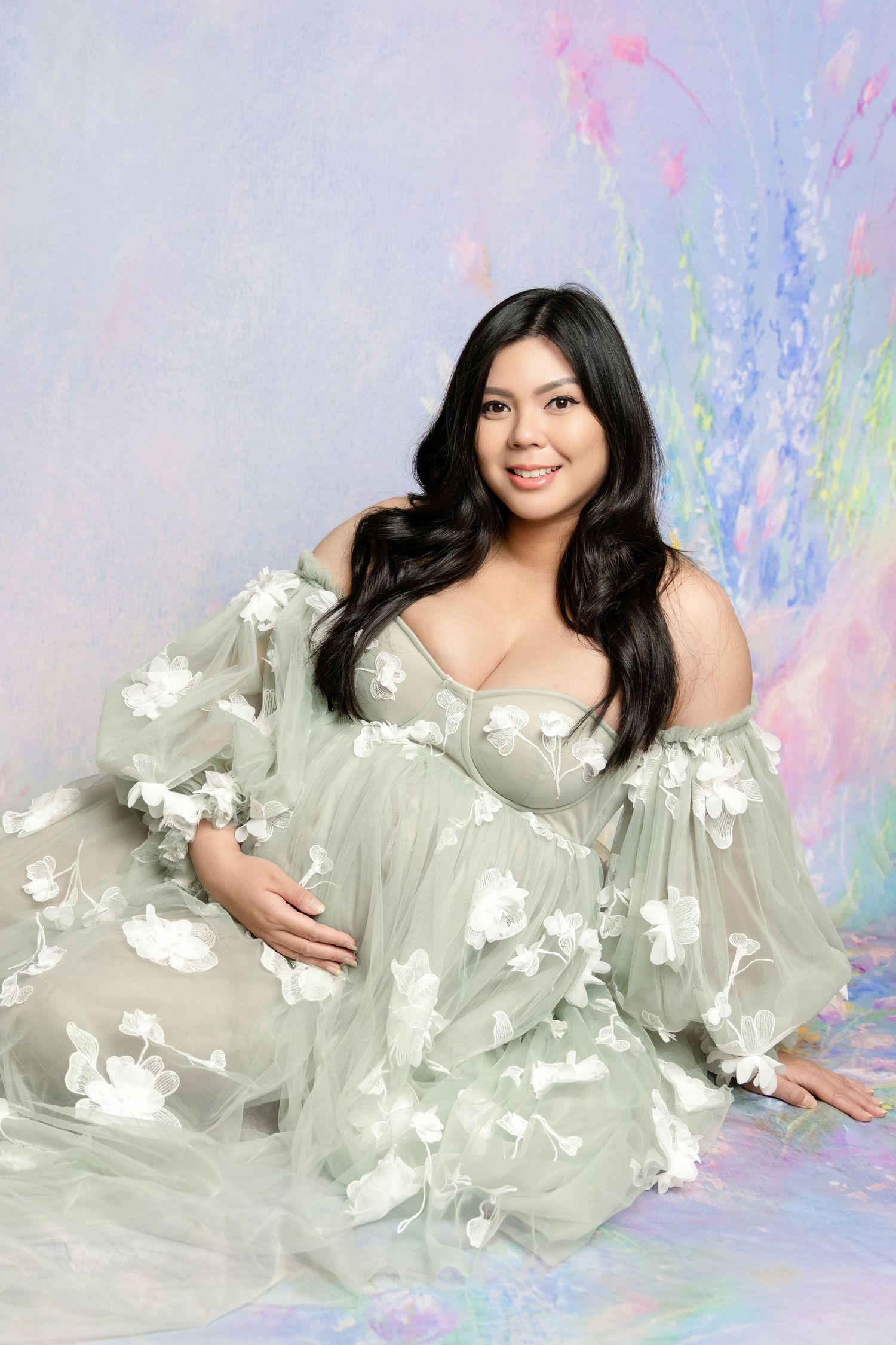 Pregnant woman in sage green floral tulle maternity gown posing gracefully against a soft pastel floral backdrop.