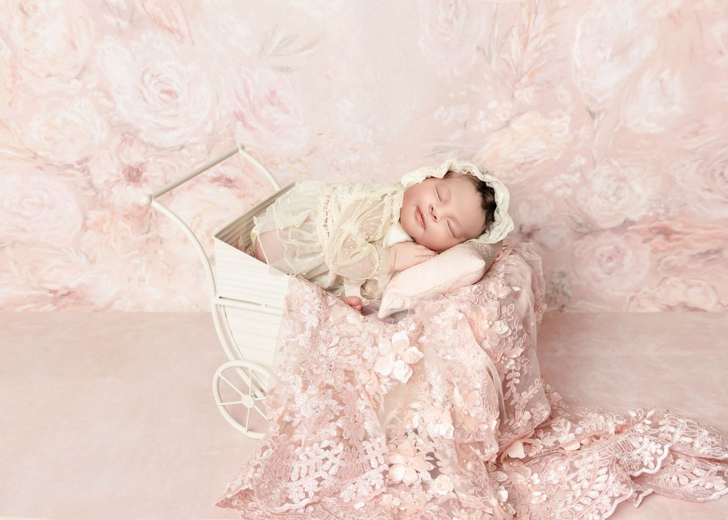 Sleeping newborn baby wrapped in cream lace in a white vintage pram on a pink floral backdrop.