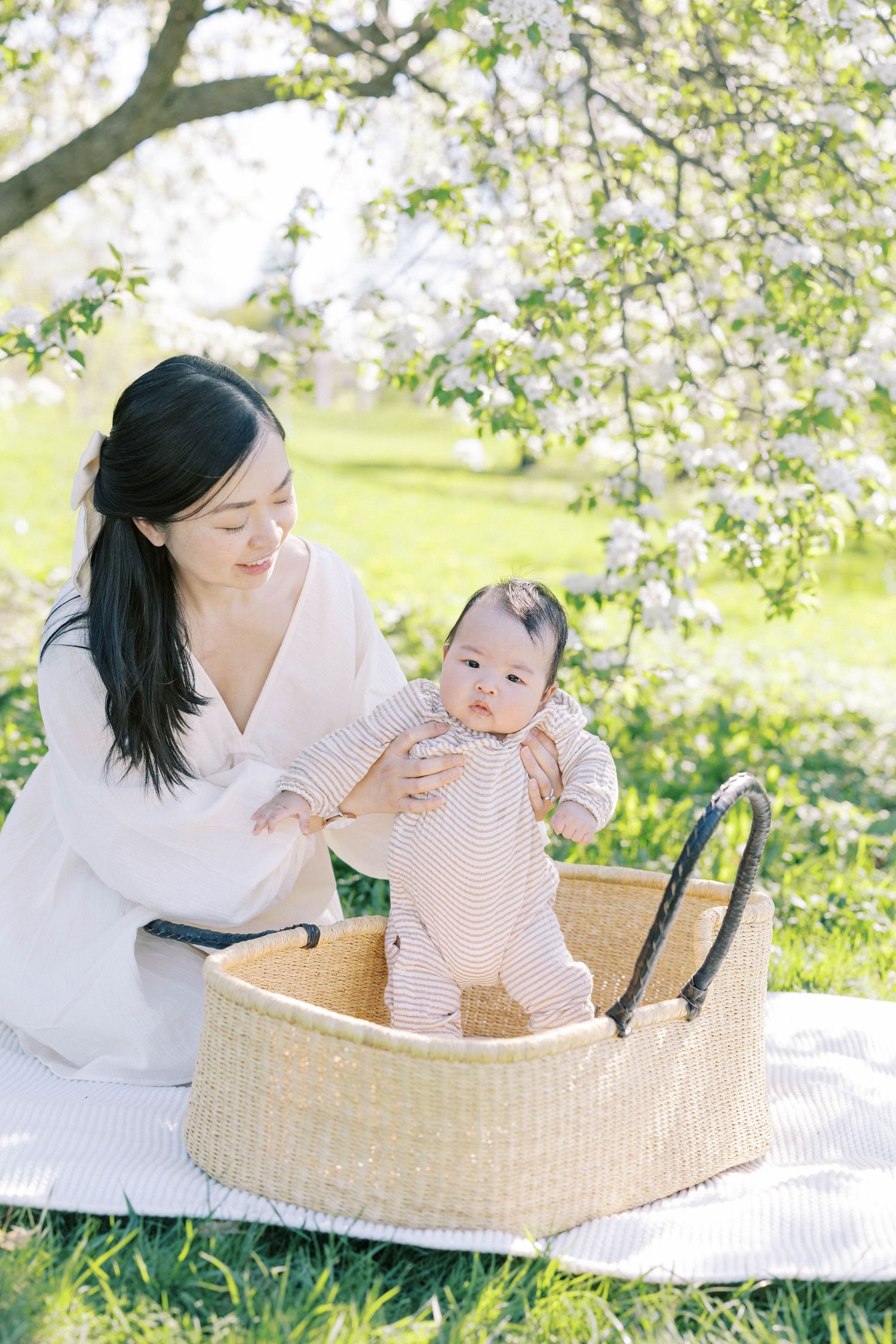 Mother in white dress helping baby stand in Moses basket on white blanket under blooming spring tree outdoors.