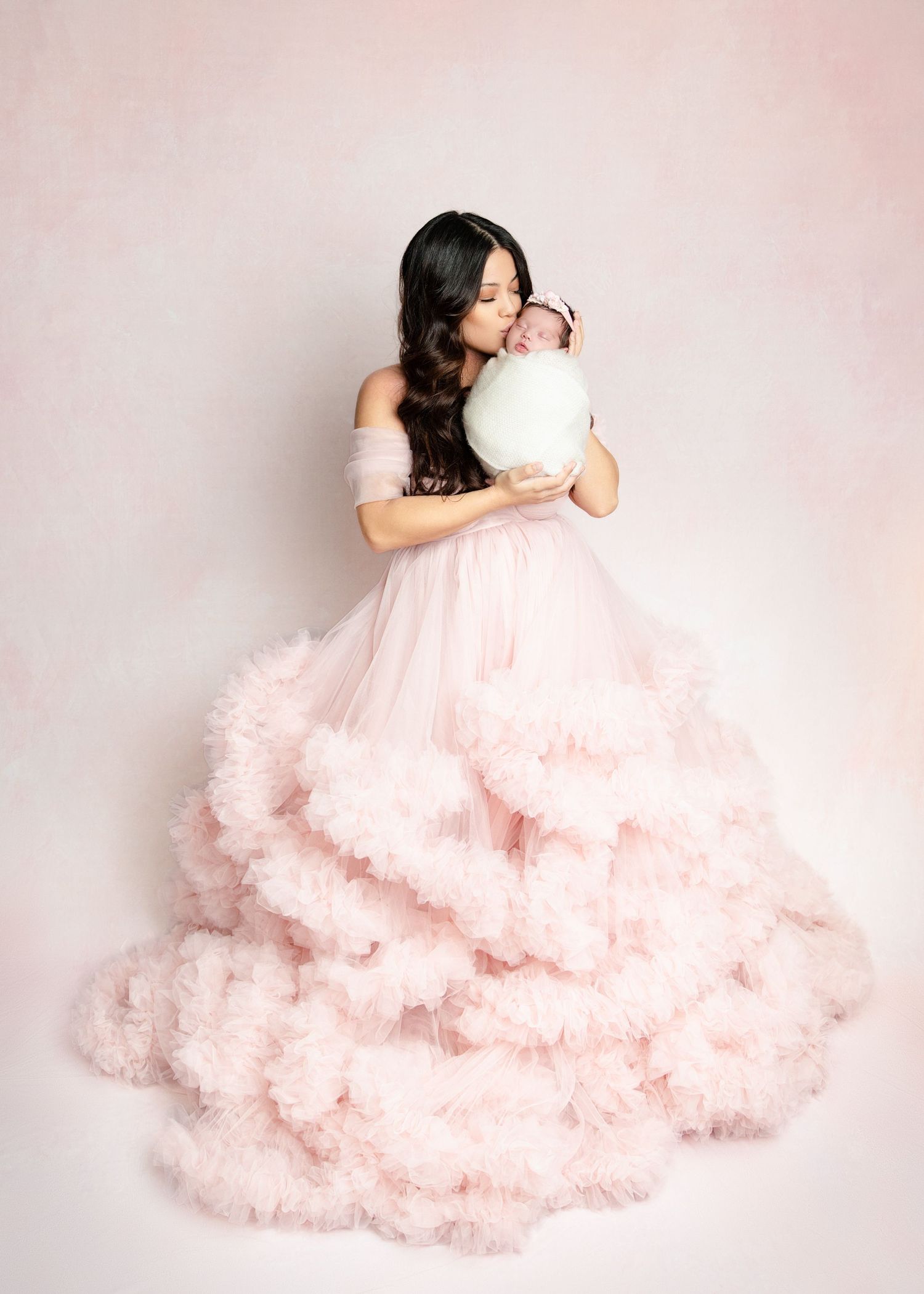 Woman in pink ruffled ballgown kissing newborn baby wrapped in mint blanket against soft pink background.