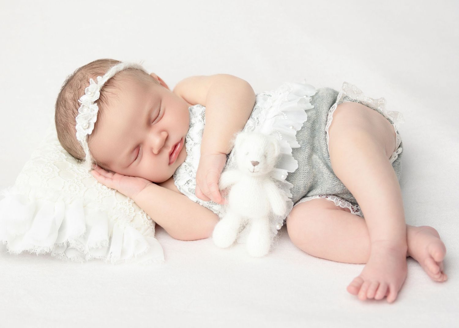 Peaceful newborn baby girl sleeping in gray ruffled outfit with white headband hugging a bunny toy.