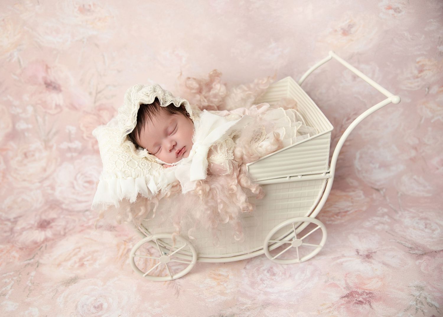 A sleeping newborn baby in a lace bonnet rests peacefully in a vintage white miniature baby carriage.