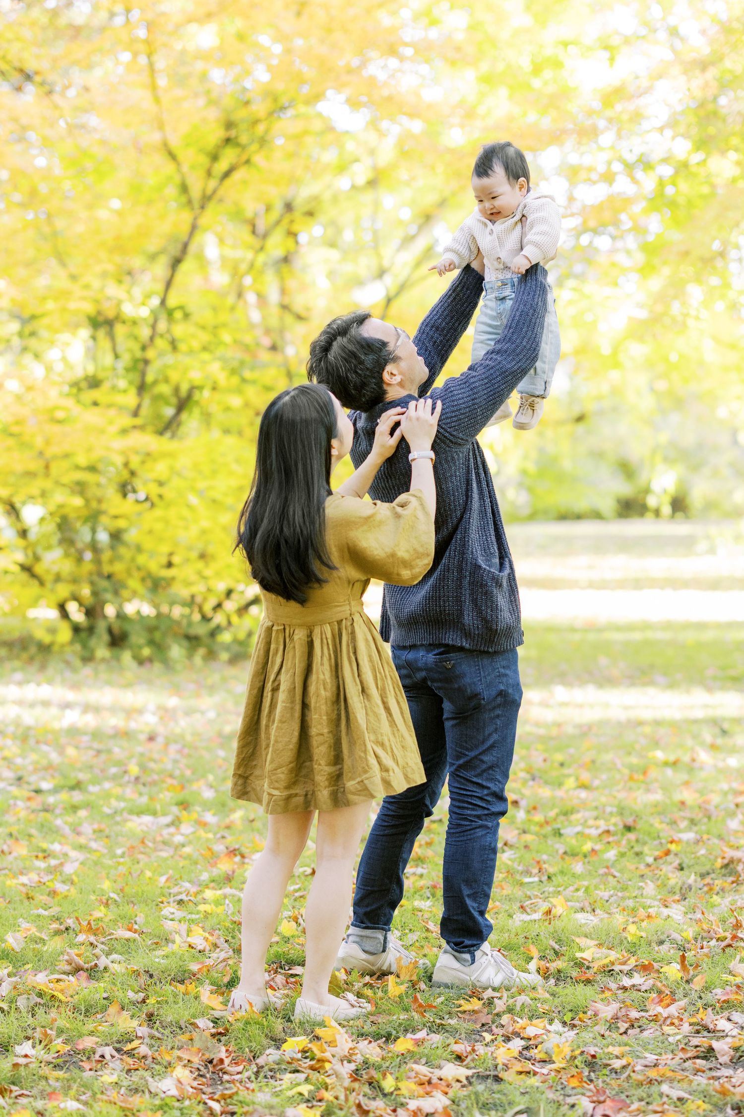Asian family lifting baby in air among colorful autumn leaves in park.