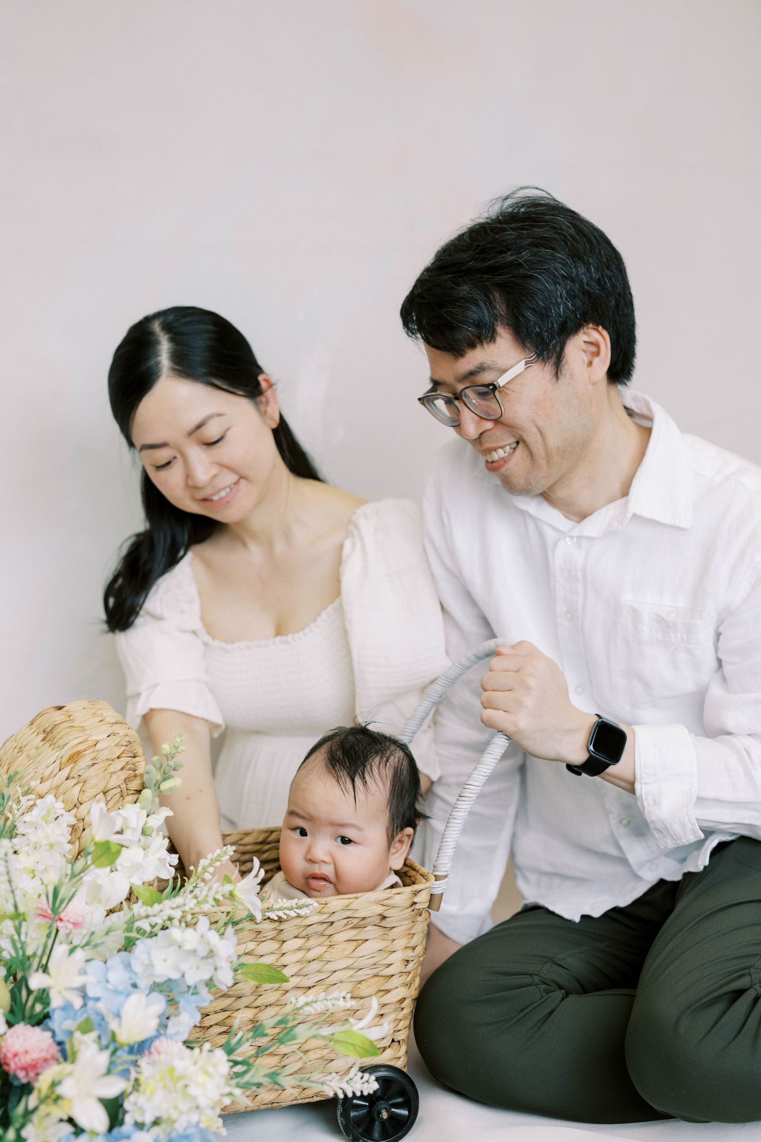 Asian family with mother, father, and baby in wicker basket surrounded by colorful flowers in studio portrait.