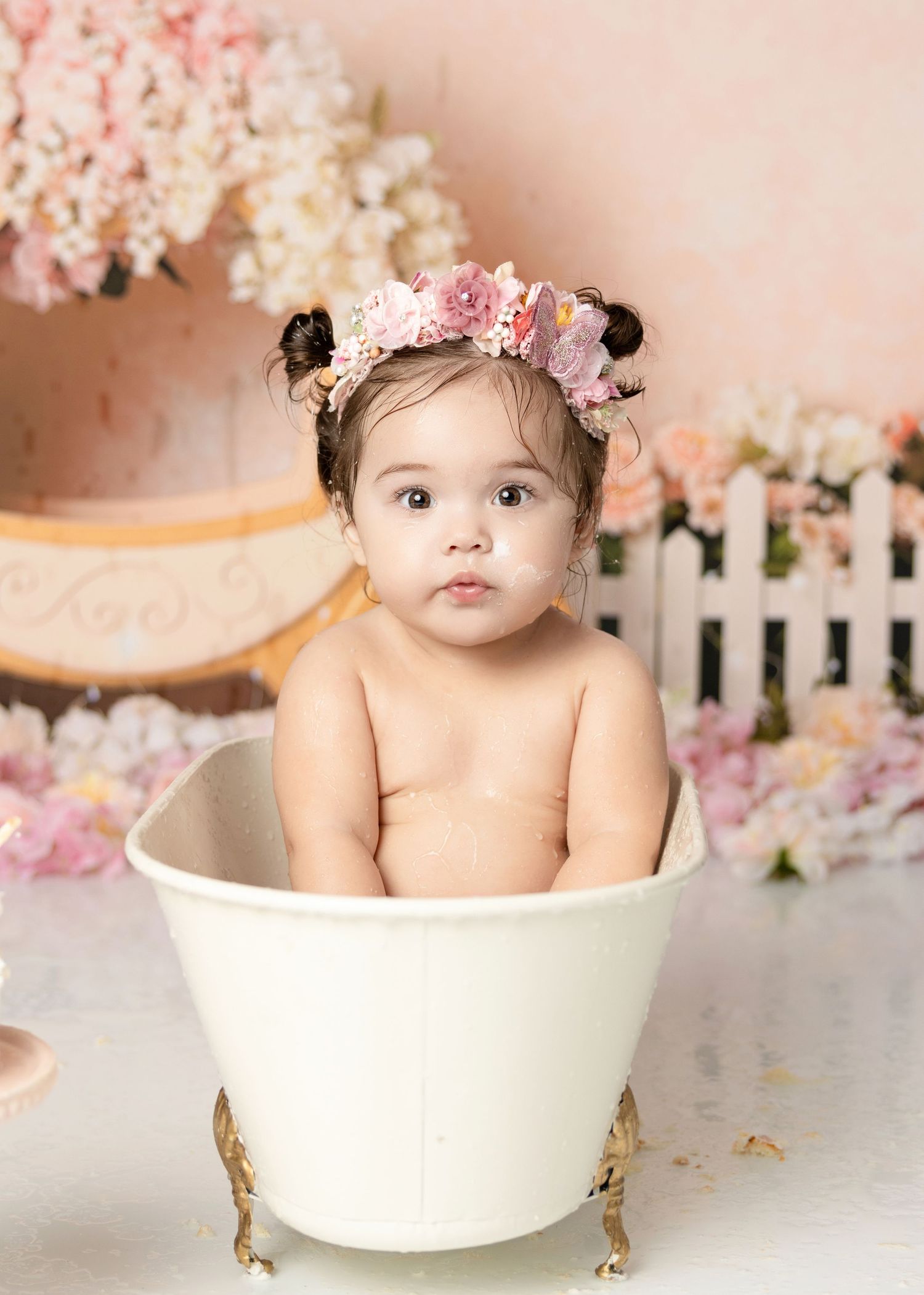 Cute baby girl with pink floral crown sits in white gold clawfoot tub during princess first birthday session.