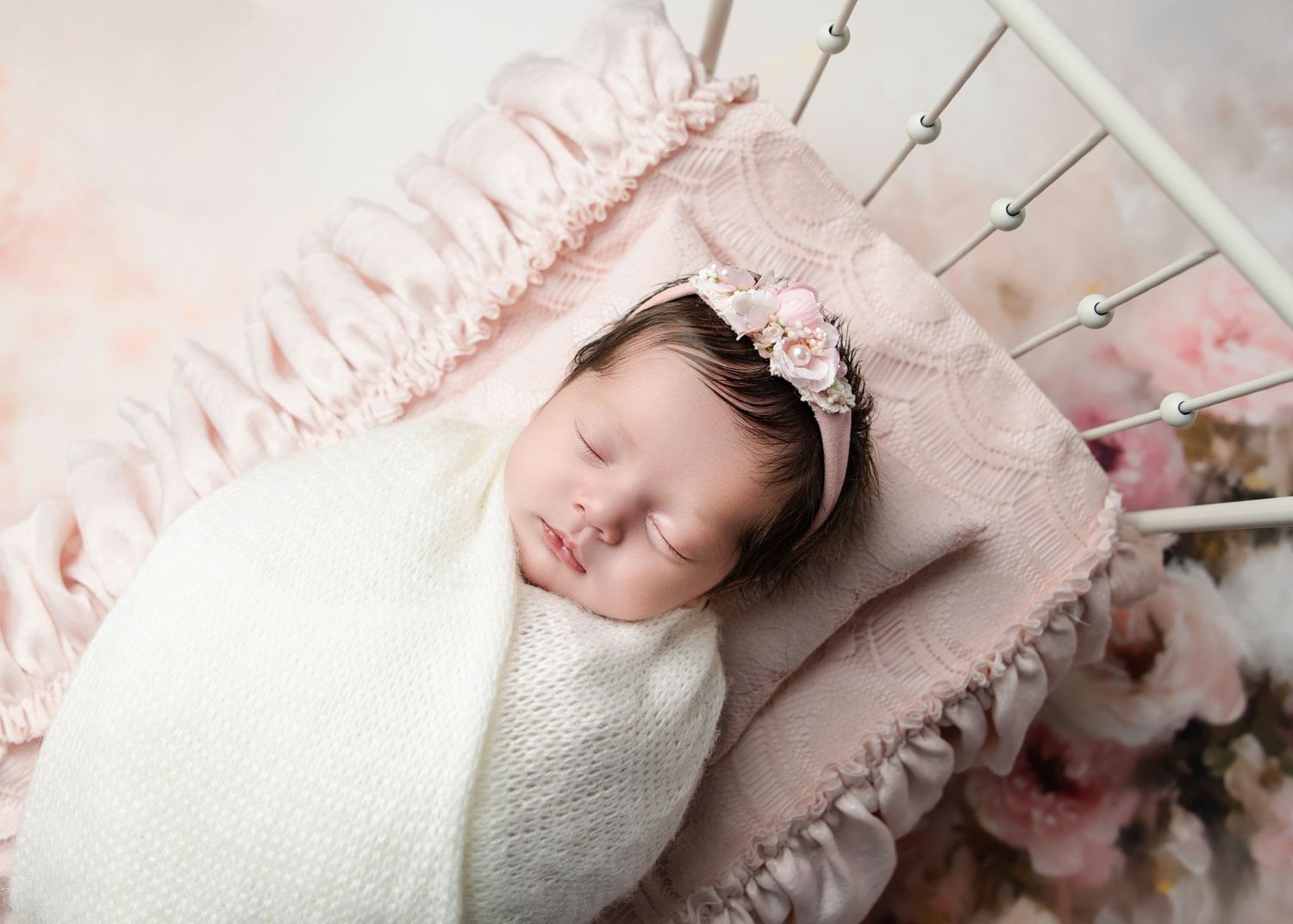 A sleeping newborn baby girl in a white wrap and floral headband rests in a vintage pink mini crib.