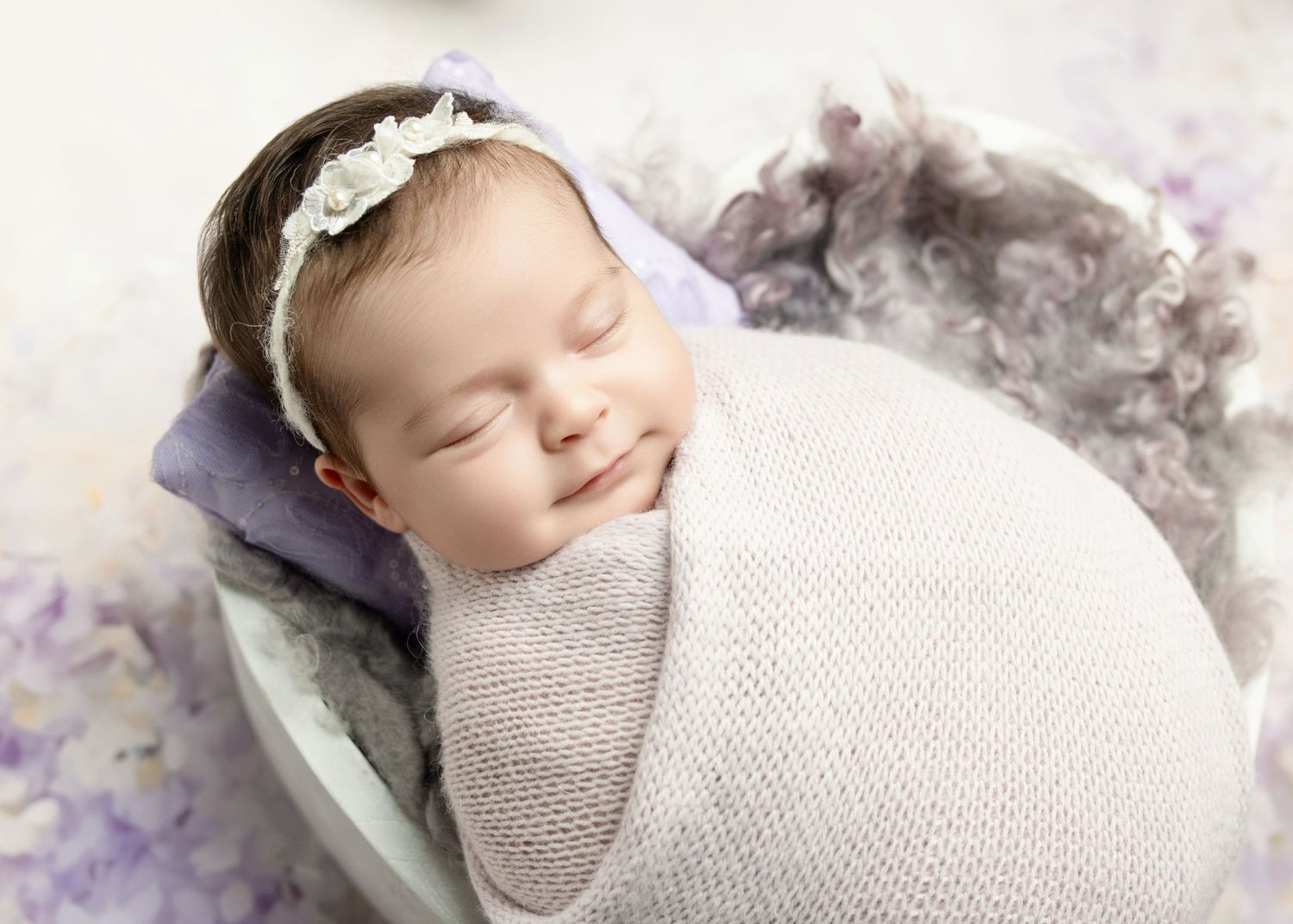 Smiling sleeping newborn in lavender knit wrap with lace headband in bowl prop.