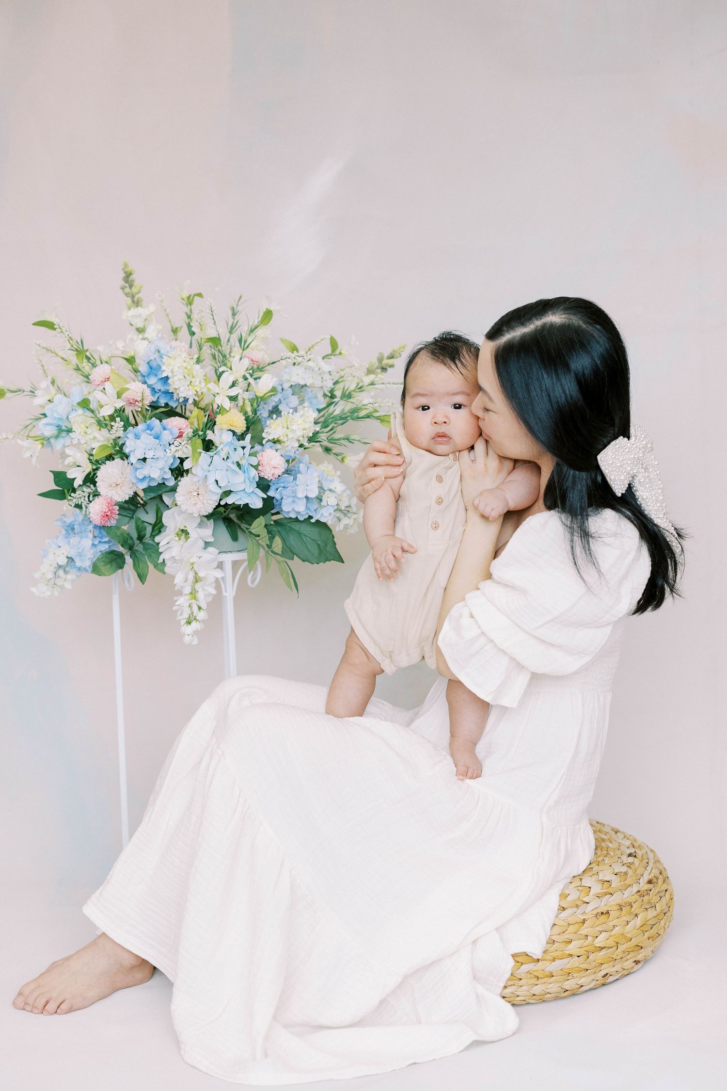 Mother in white dress kissing her baby while sitting on a wicker pouf beside blue and pink floral arrangement.