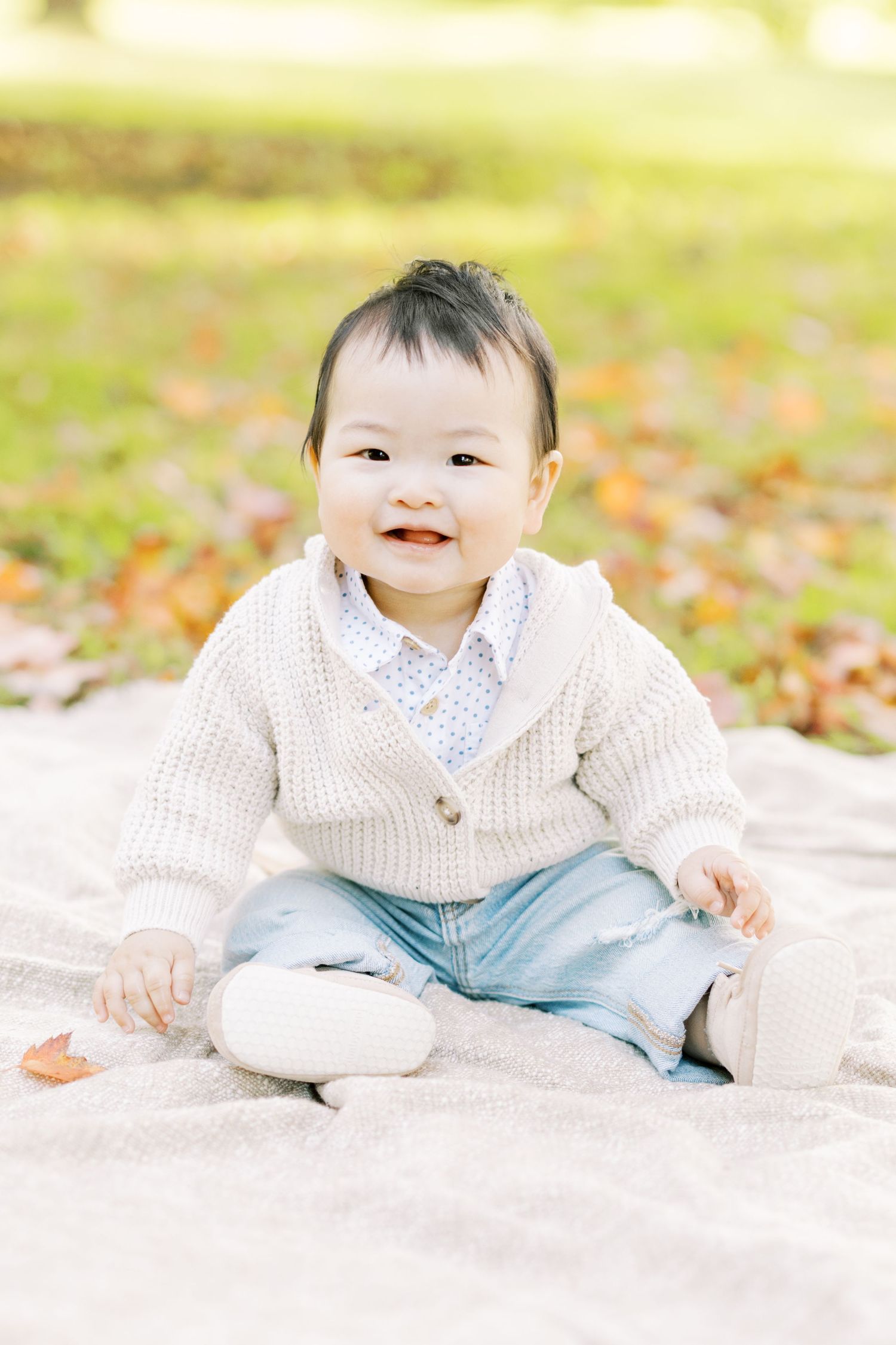 An adorable baby in a cream sweater sits smiling on a blanket among autumn leaves.