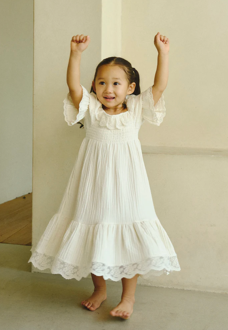 A joyful toddler girl in a white lace dress raises her arms in celebration, smiling brightly while standing barefoot.