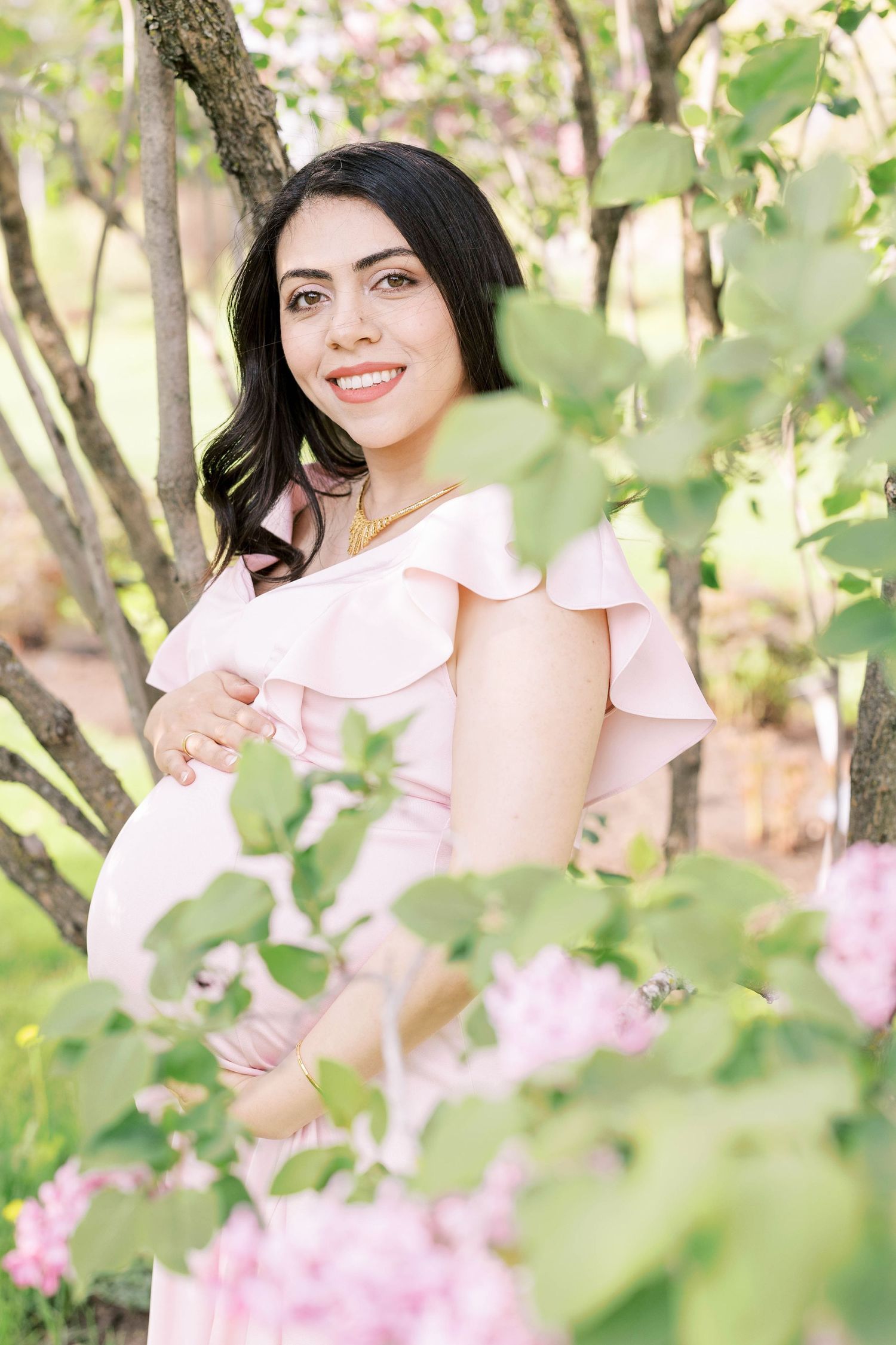 Smiling pregnant woman in pink ruffled dress posing among blooming spring trees and flowers.
