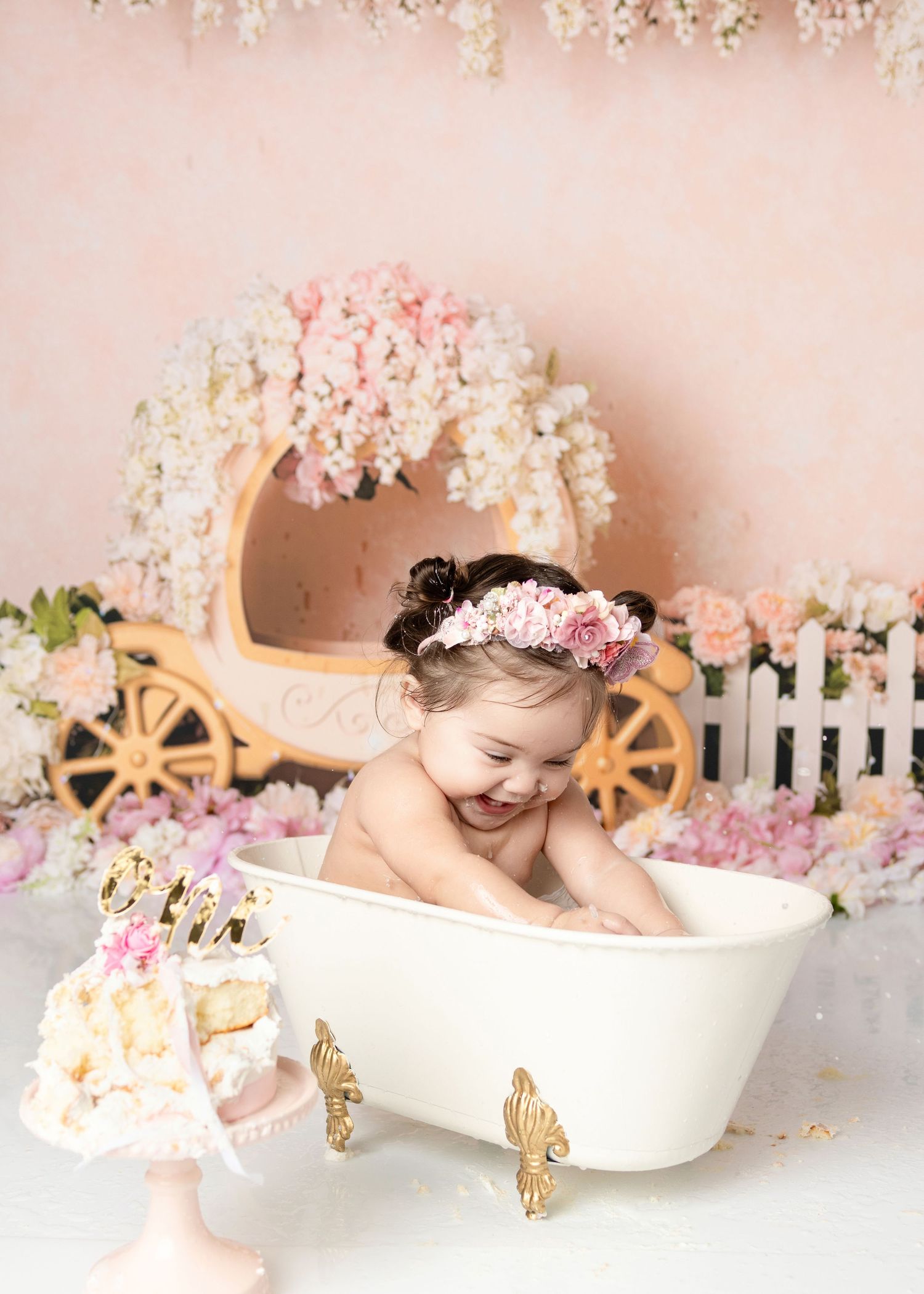 Giggling baby girl in floral headband plays in white clawfoot tub with princess carriage and flowers backdrop.