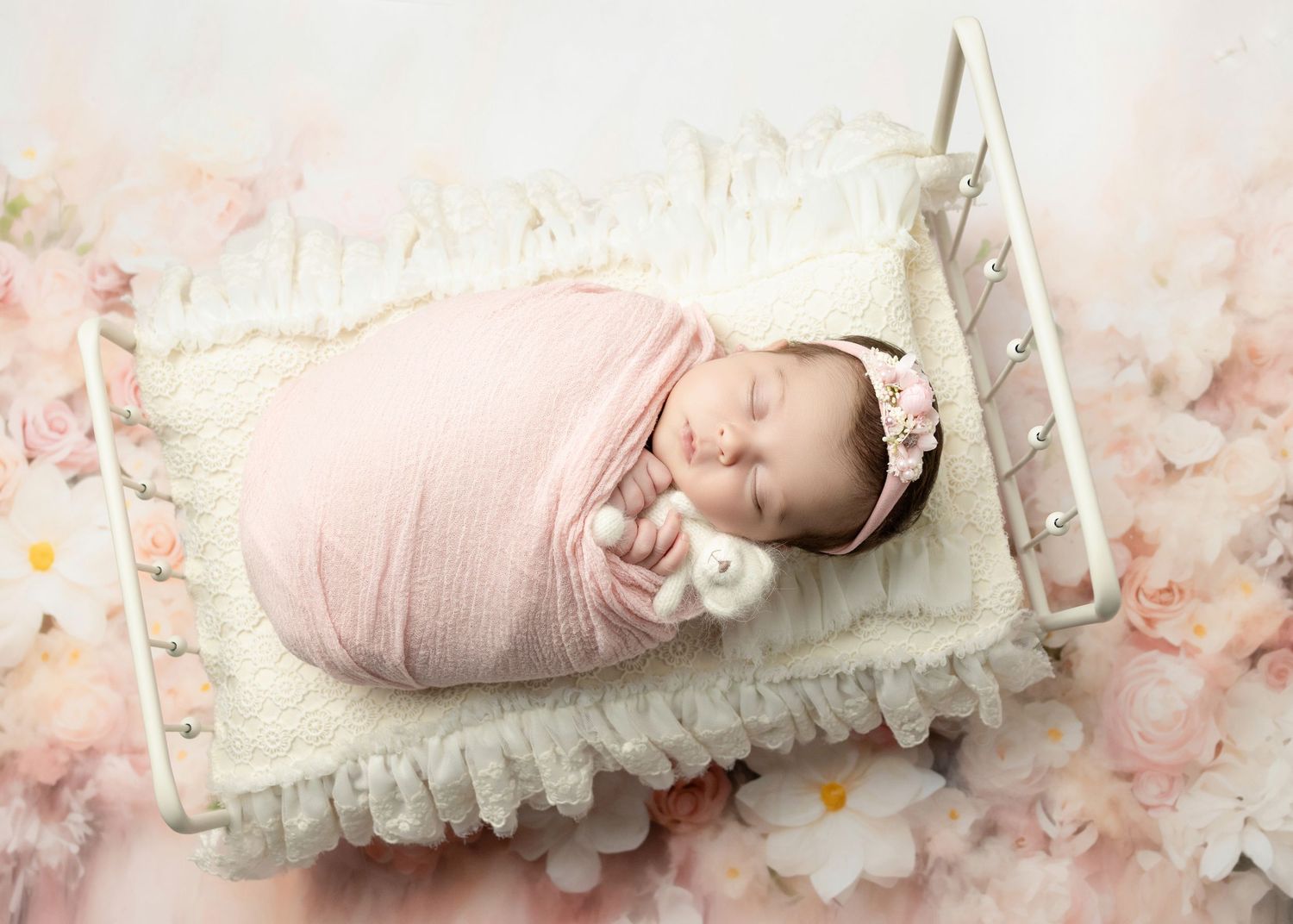 Newborn baby in pink swaddle with floral headband sleeping on miniature white bed.