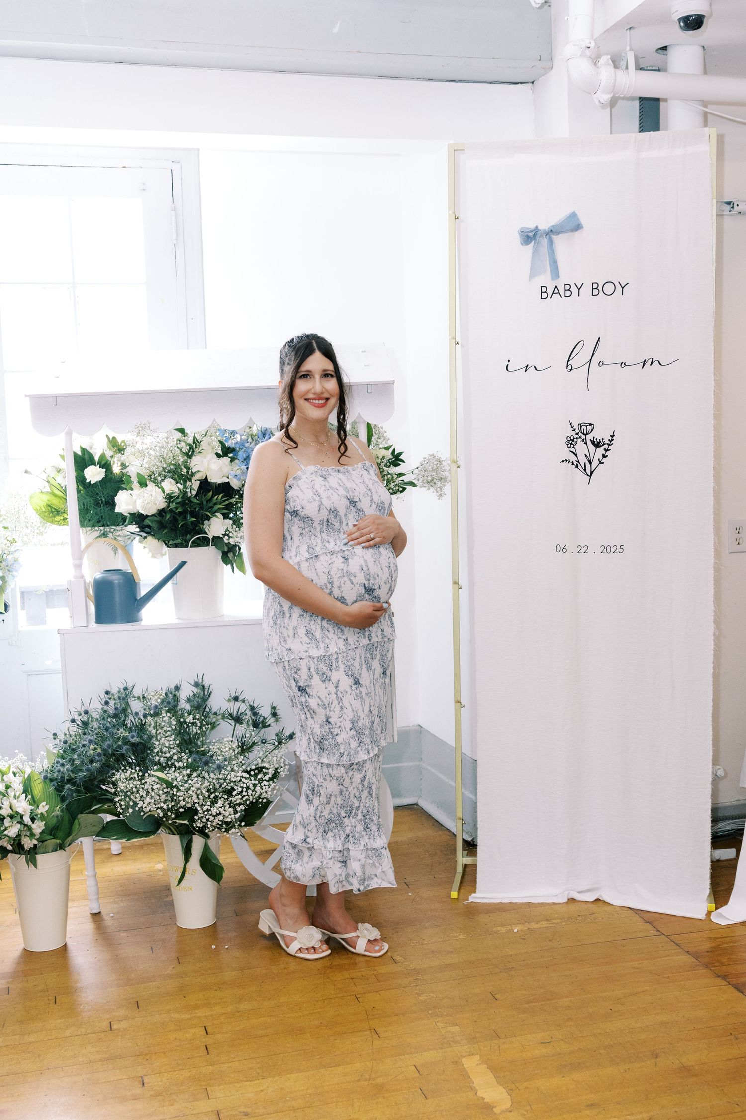Pregnant woman in floral dress poses beside a Baby Boy in Bloom baby shower welcome sign with flowers.