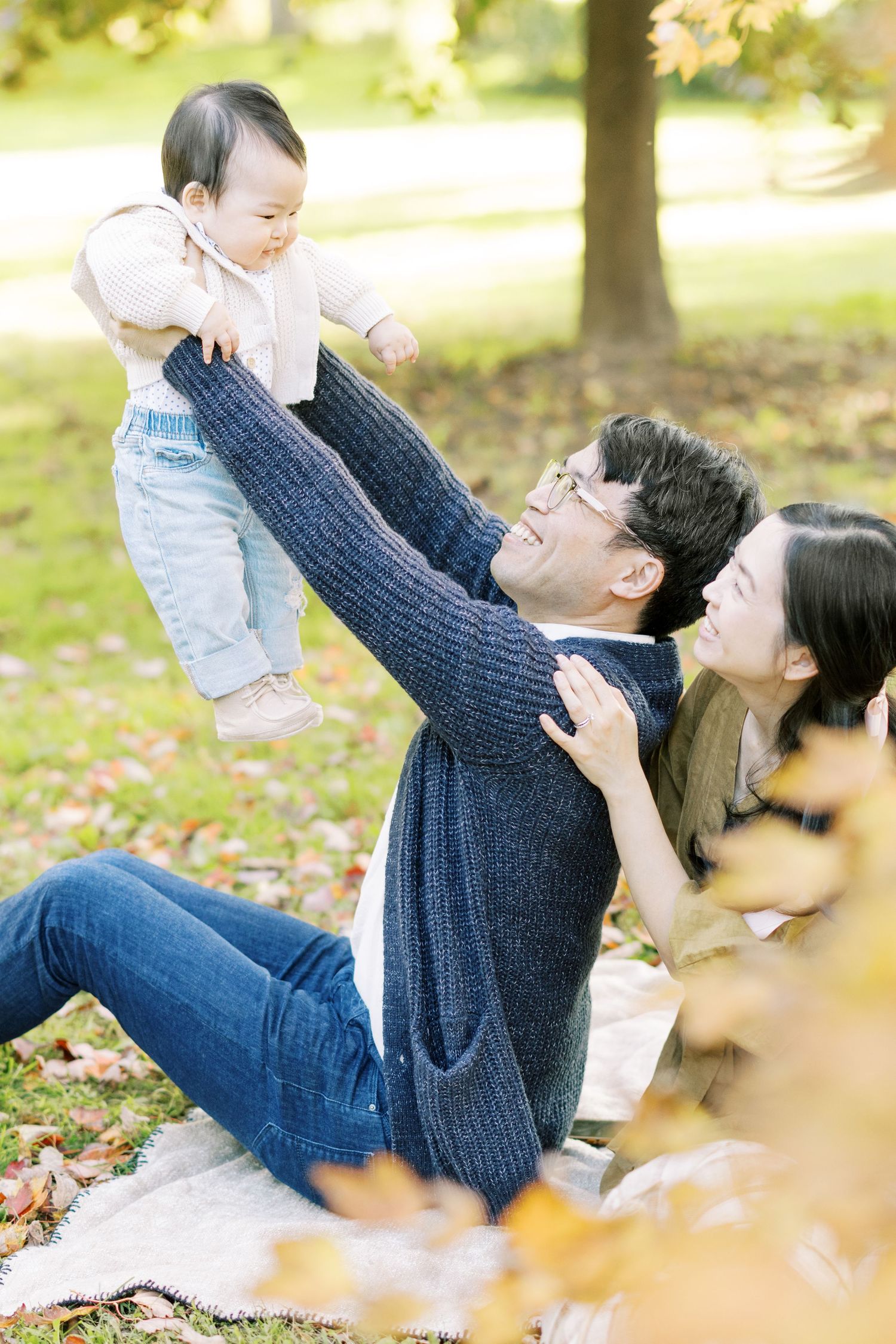 A father lifts his laughing baby in the air while sitting on a blanket in an autumn park.